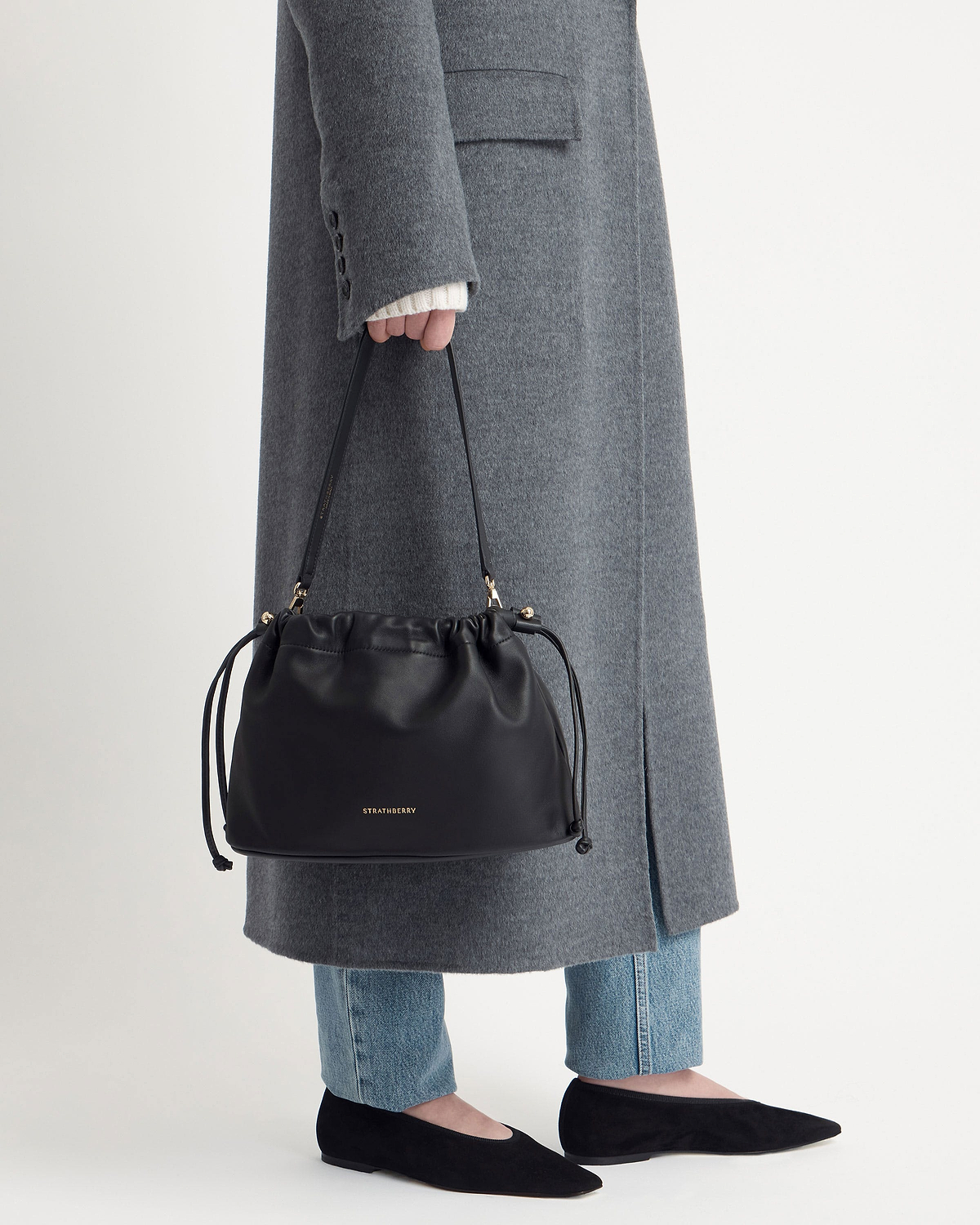 Black Strathberry Charlotte Midi drawstring bag held by model wearing grey coat.
