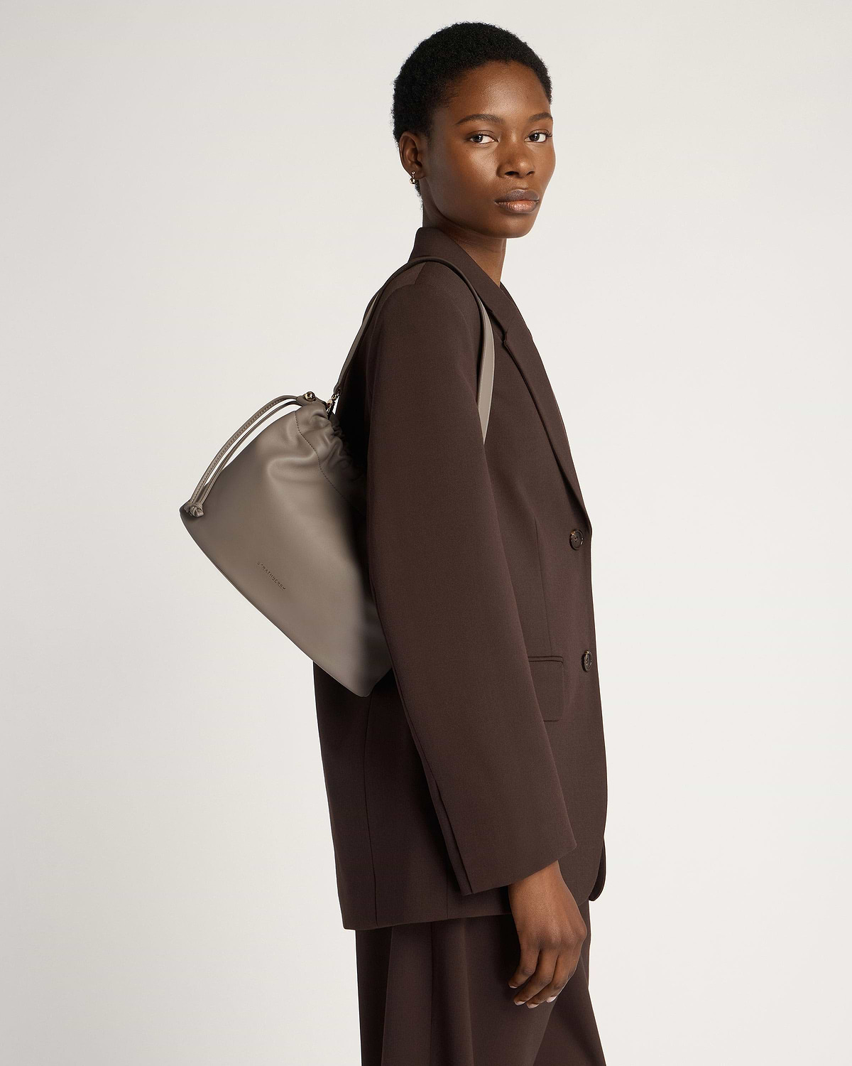 Model wearing a brown blazer carrying a taupe drawstring shoulder bag.