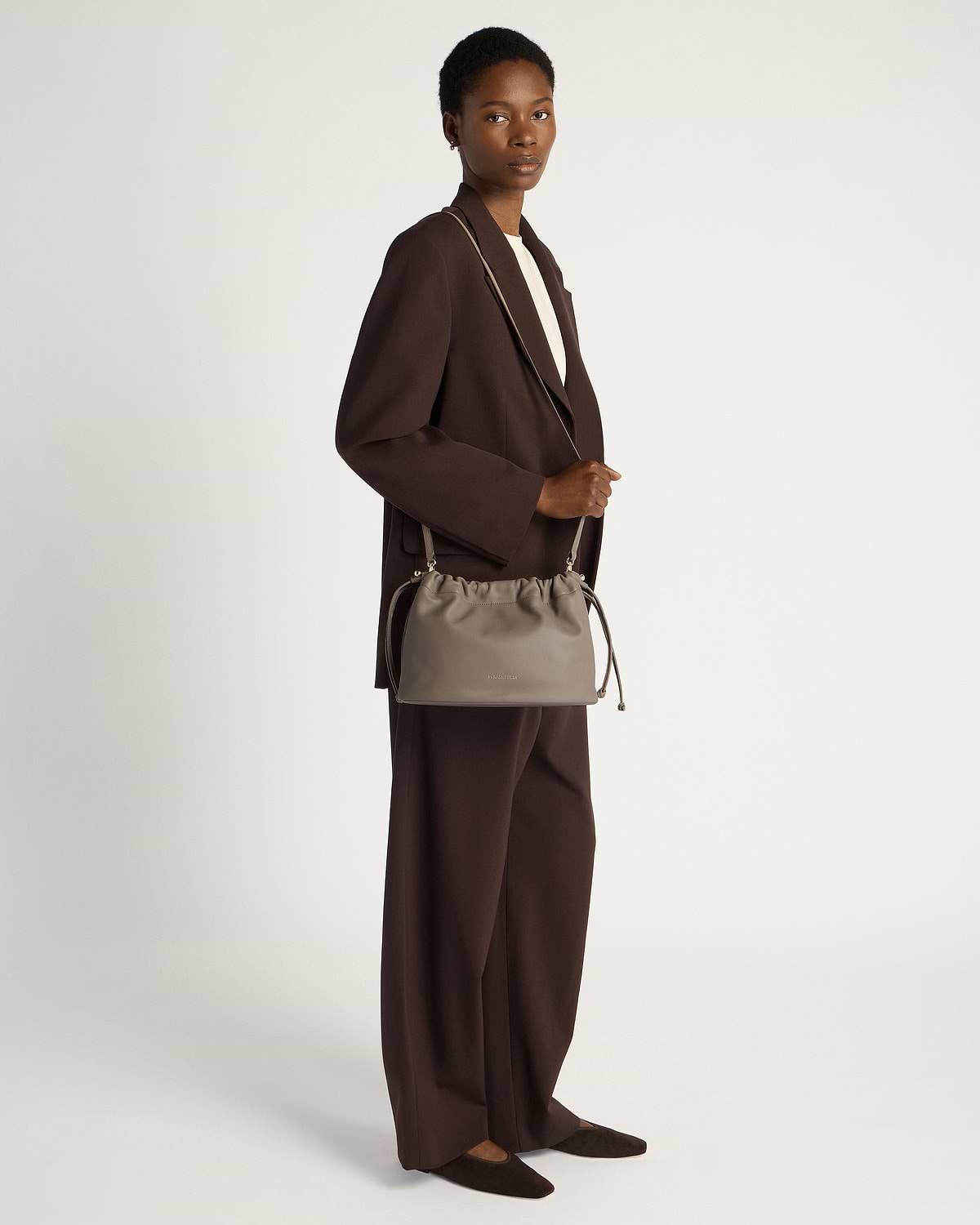 Model in brown suit holding a taupe drawstring shoulder bag at her side.