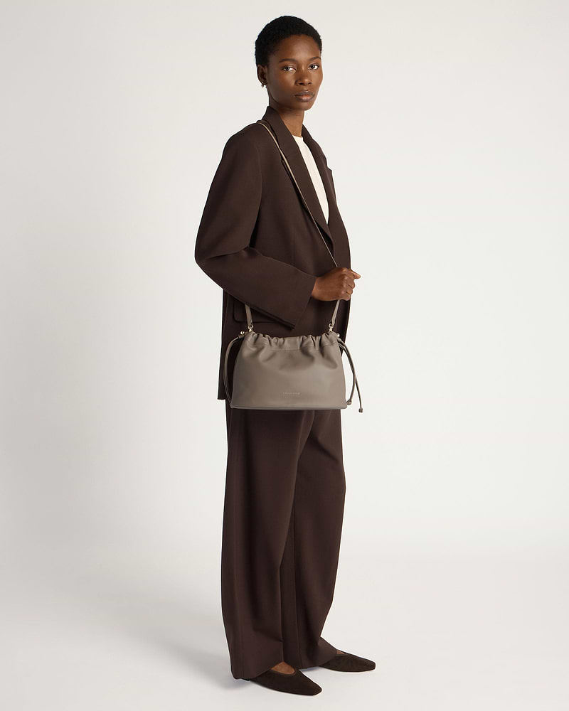 Model in brown suit holding a taupe drawstring shoulder bag at her side.