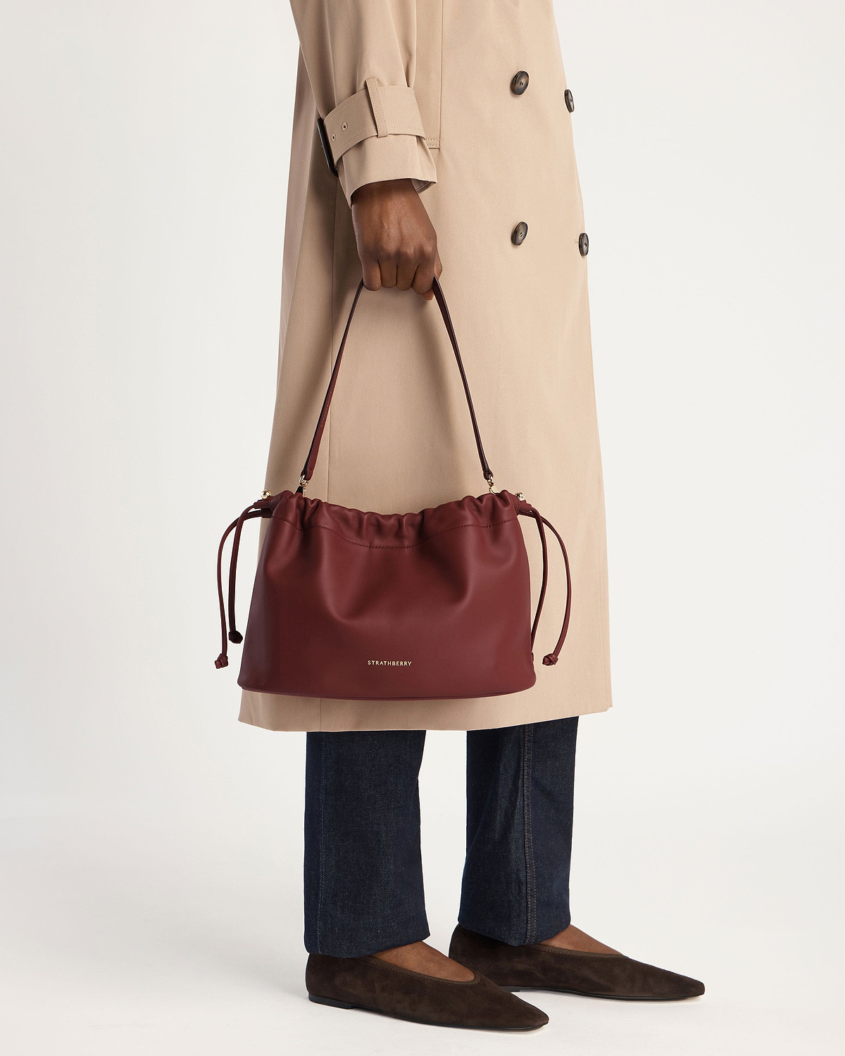 Model holds an oxblood Charlotte Midi drawstring bag with Strathberry logo.