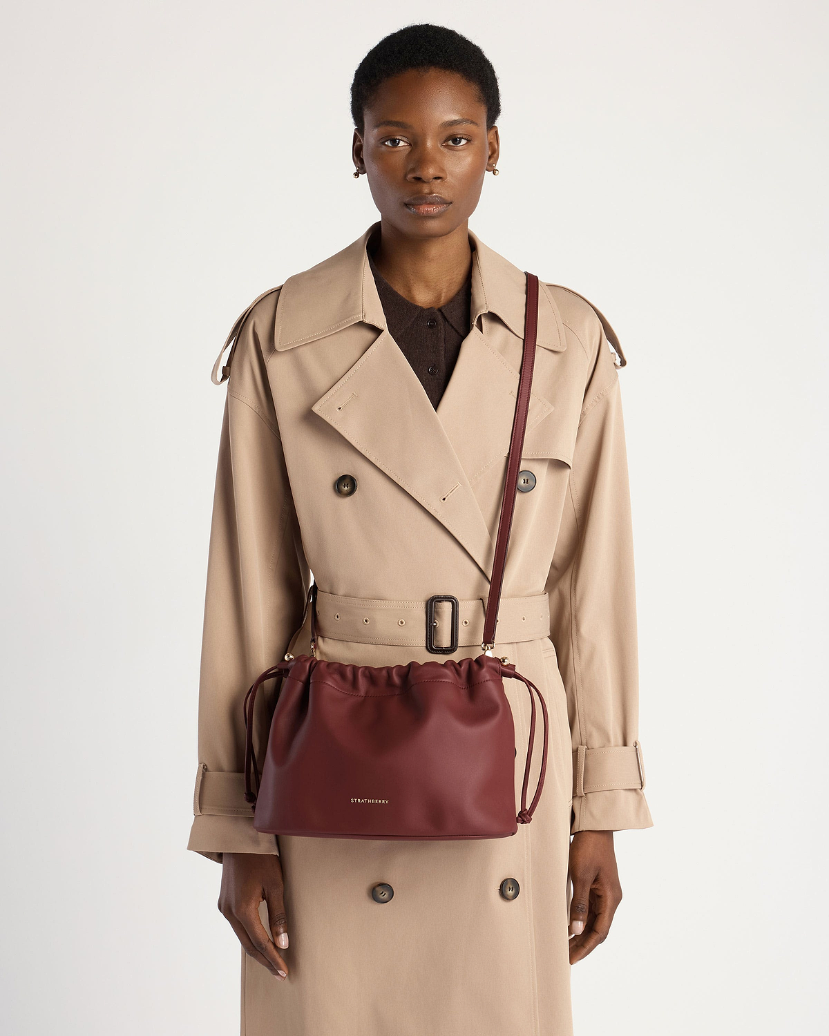 Model wearing beige trench coat carrying oxblood drawstring crossbody bag with Strathberry logo.