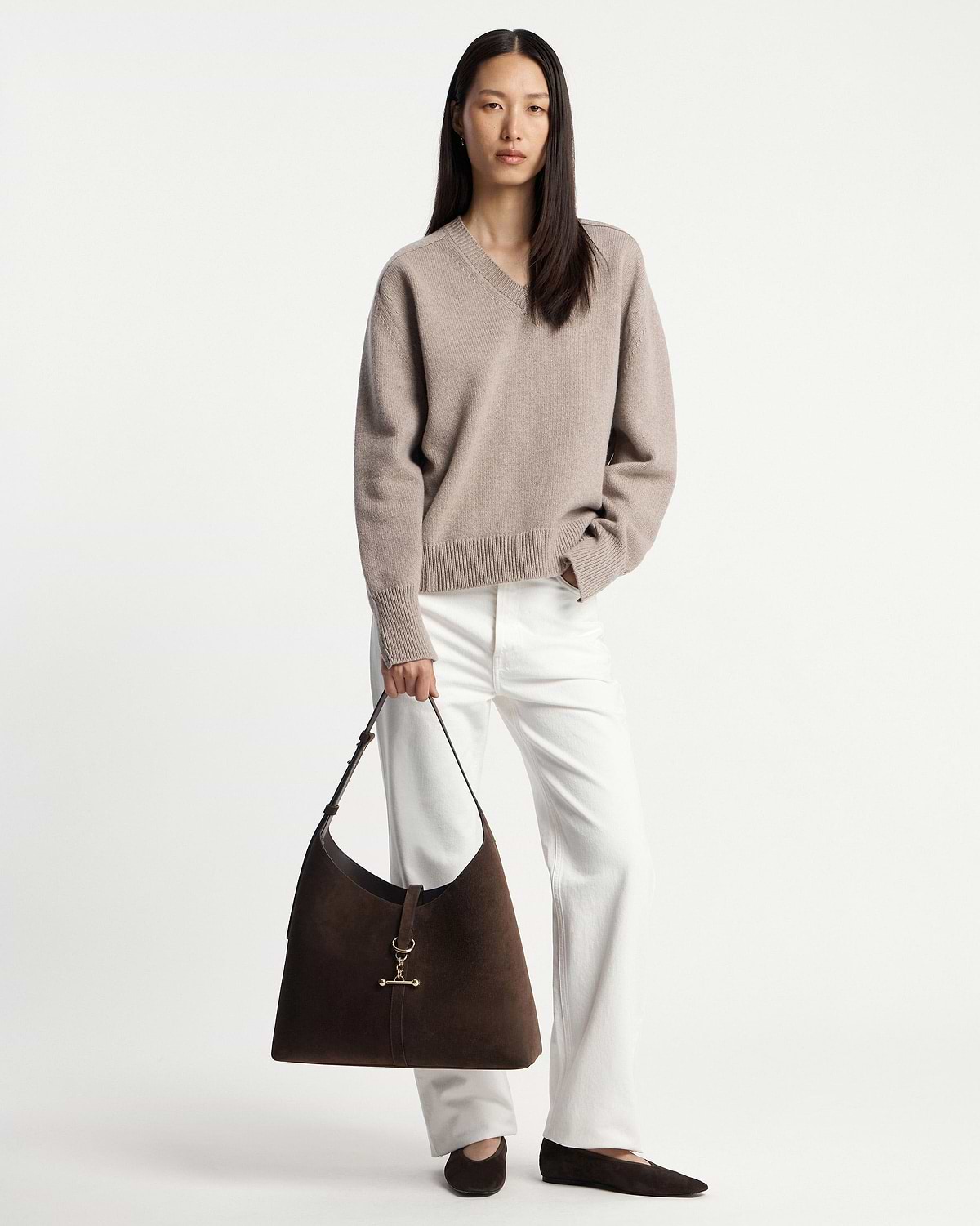 Model wearing beige V‑neck jumper and white trousers holding chocolate suede hobo bag.