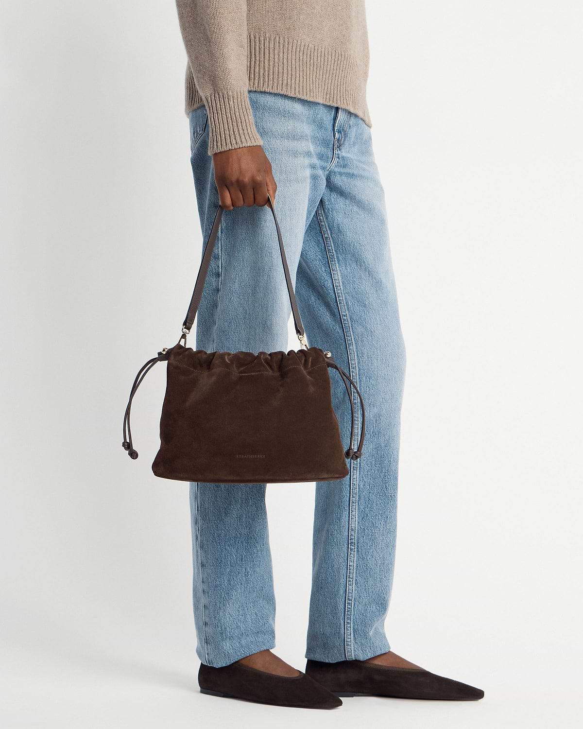 Chocolate suede Strathberry Charlotte midi drawstring bag held by model wearing jeans and flats.