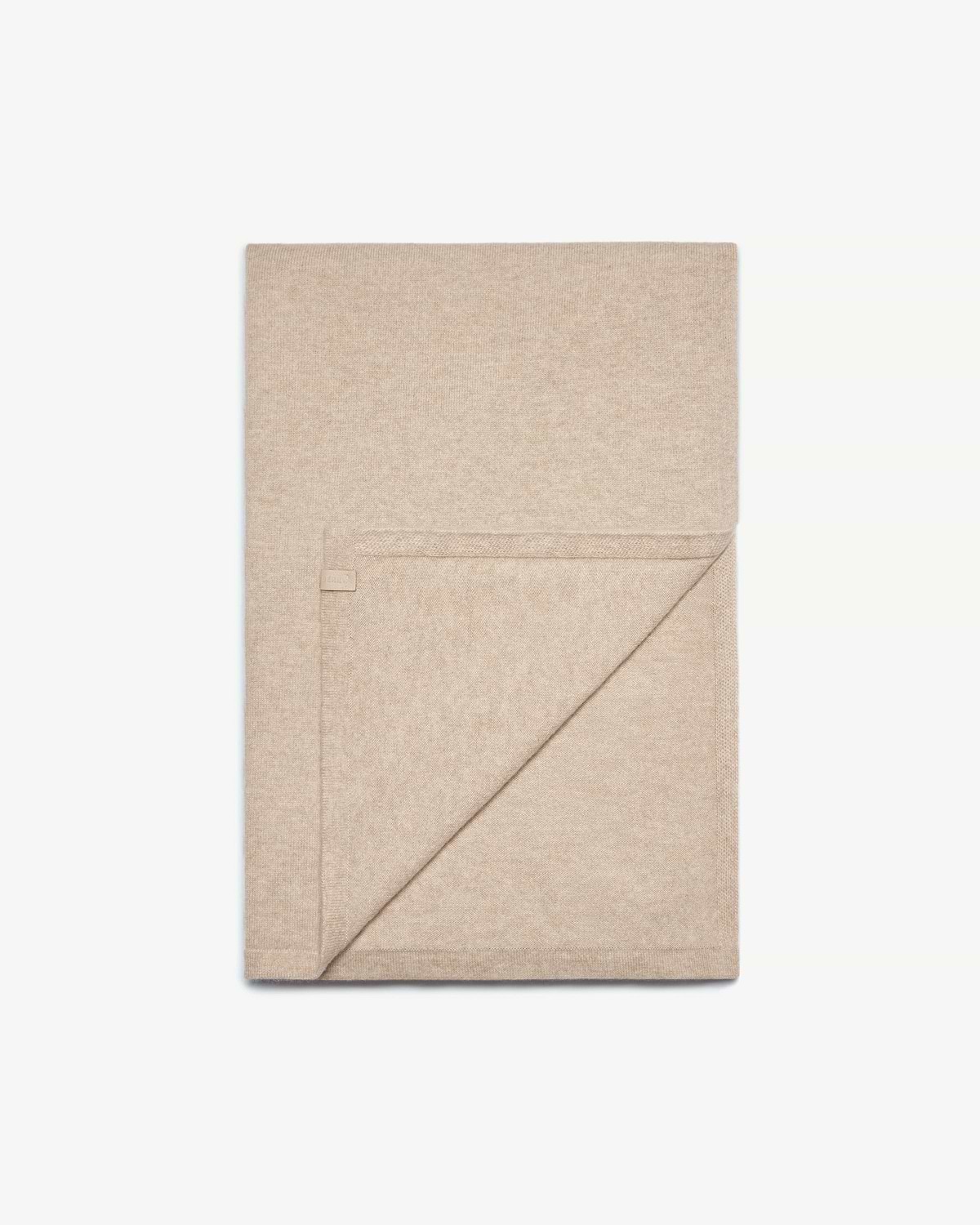 Folded oat-coloured cashmere travel wrap with a stitched corner tag and folded edge