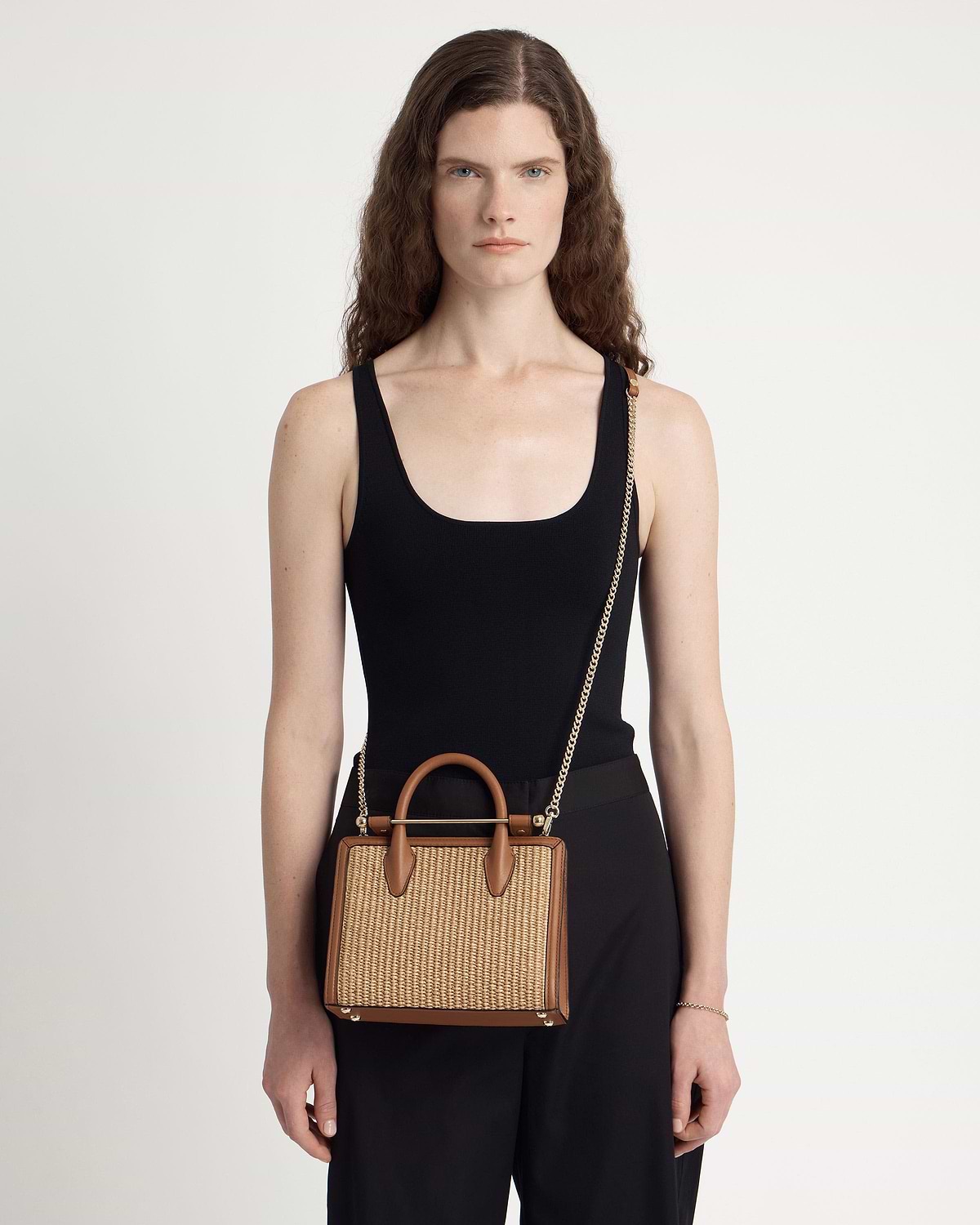 Model wearing black top holding tan and natural raffia mini tote with chain strap.