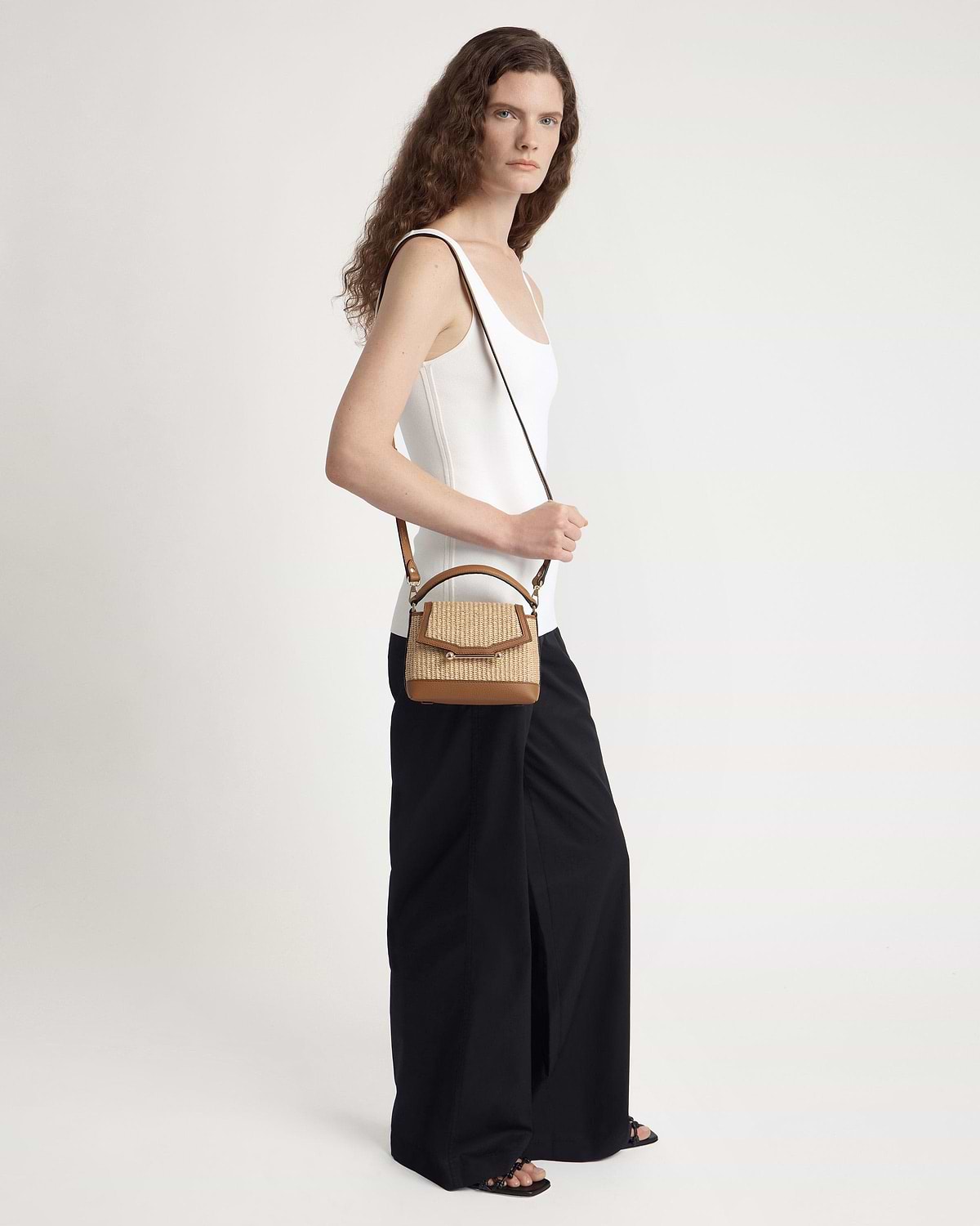Model wearing white vest top and black wide trousers carrying tan raffia crossbody bag.