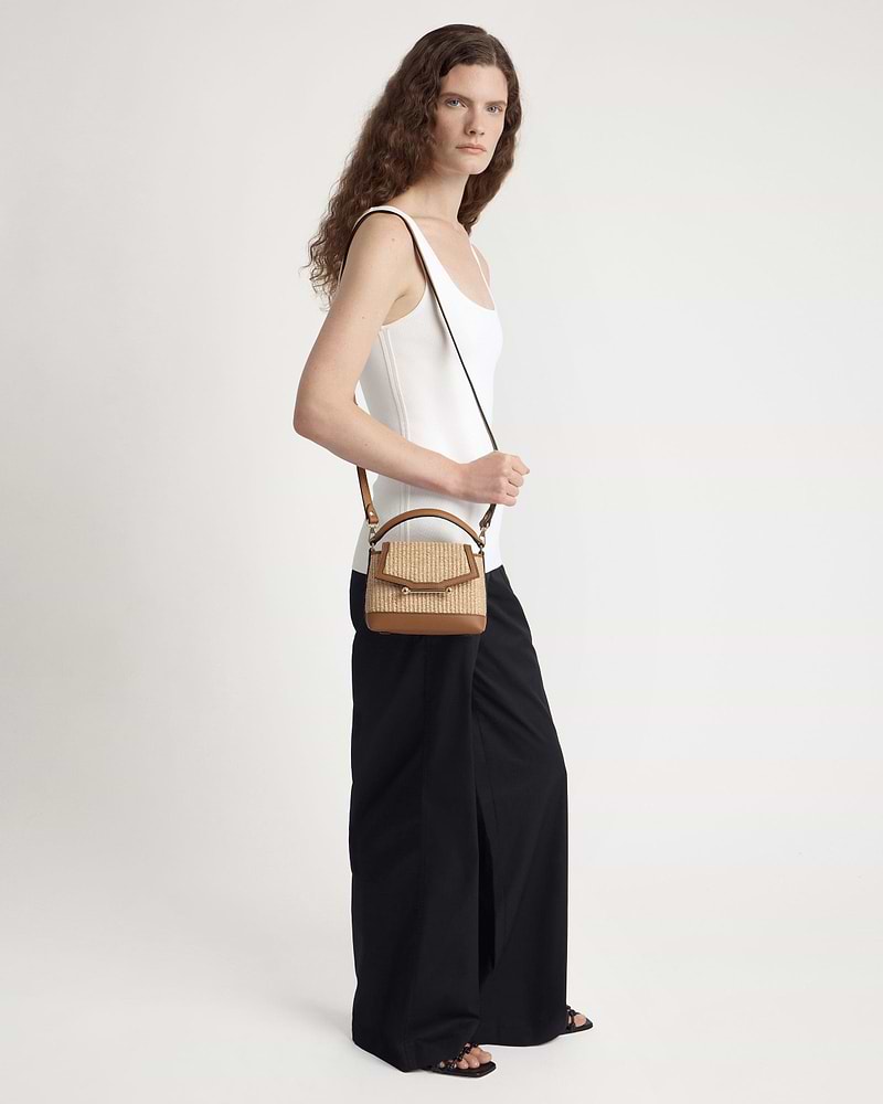 Model wearing white vest top and black wide trousers carrying tan raffia crossbody bag.