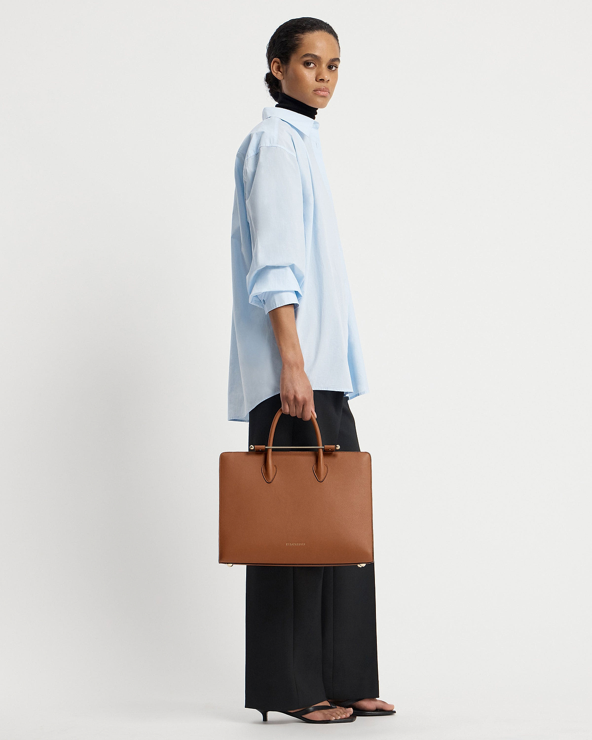 Model holding chestnut leather Strathberry tote with top handles and gold bar.