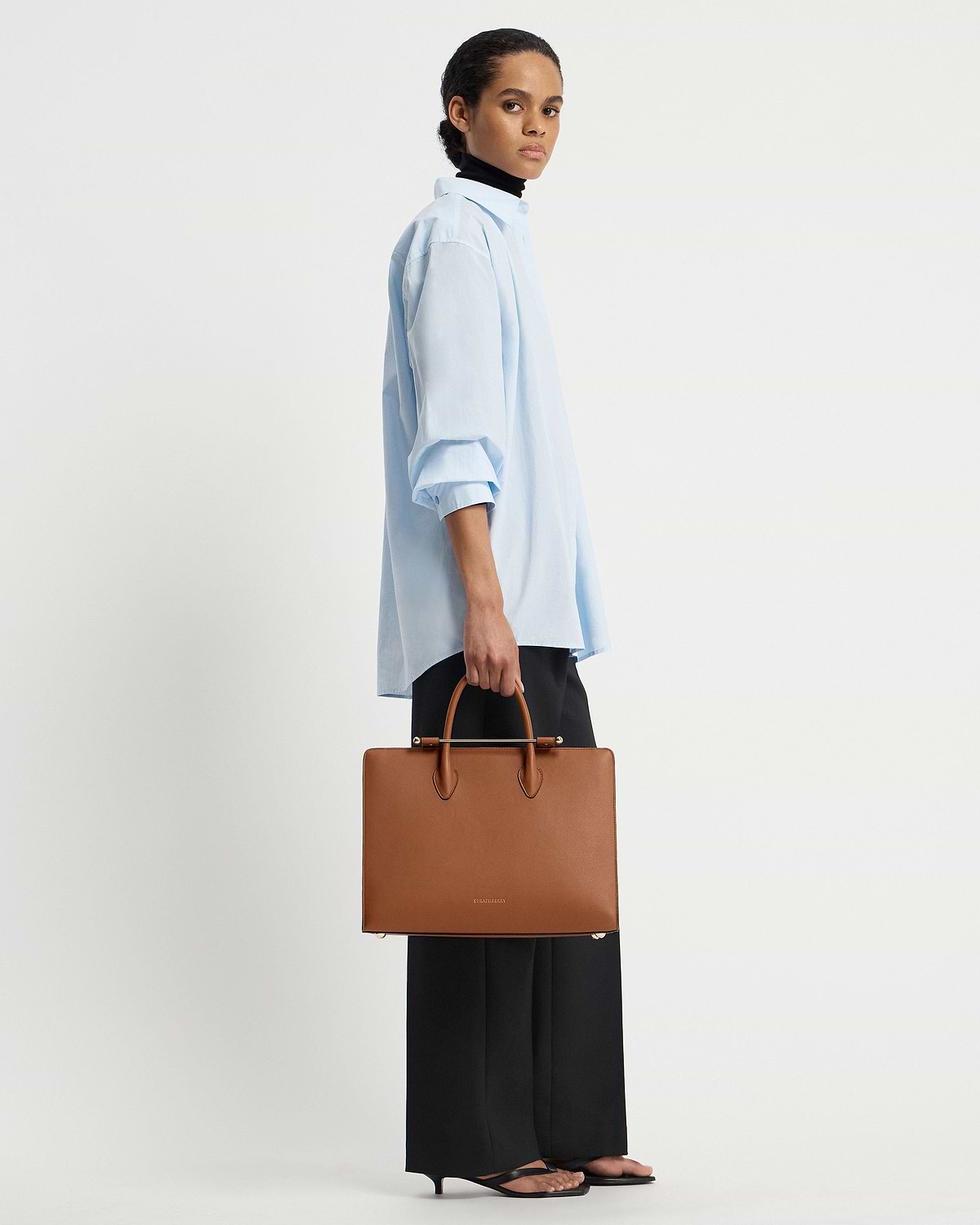 Model holding chestnut leather Strathberry tote with top handles and gold bar.