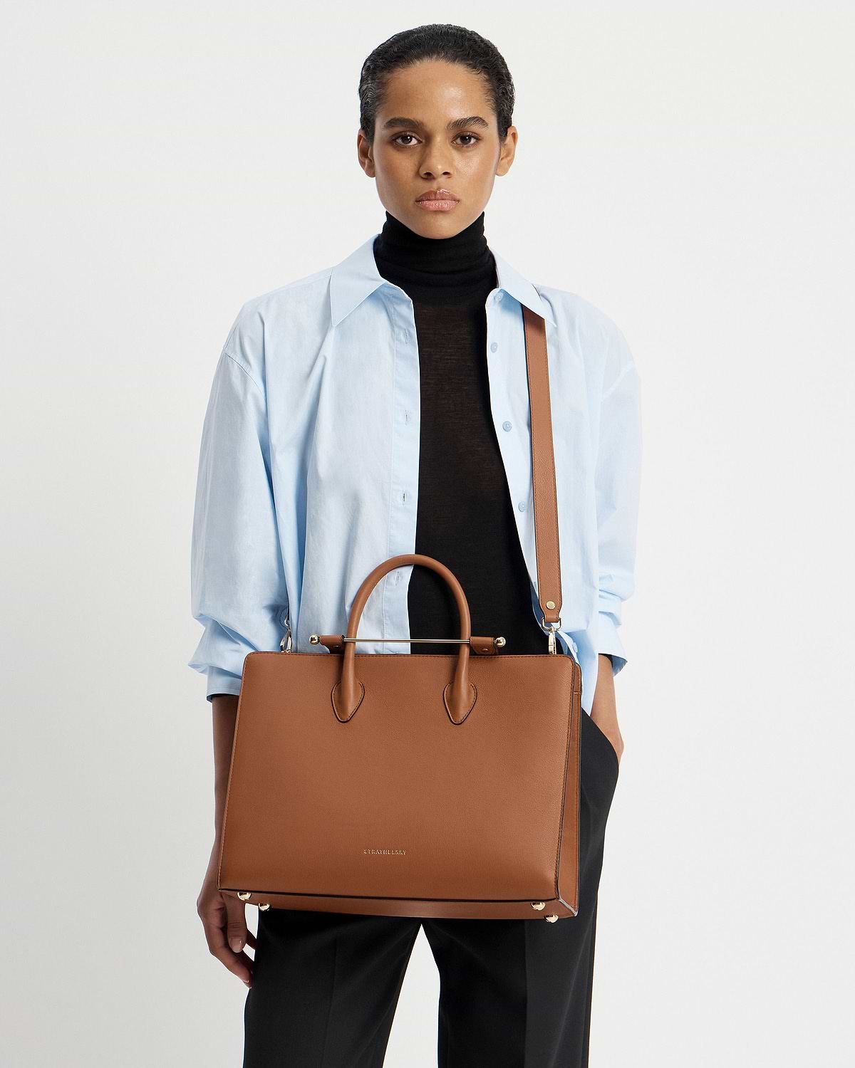Model wearing light blue shirt and black turtleneck holding Strathberry chestnut tote.