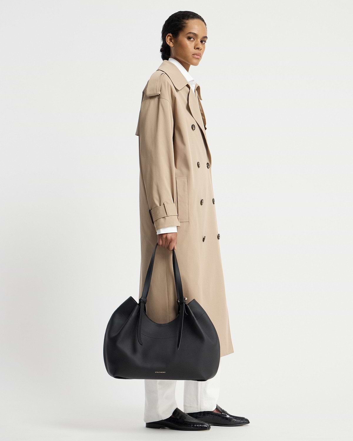 Model in a beige trench coat holding a black Strathberry Barra Tote at her side.