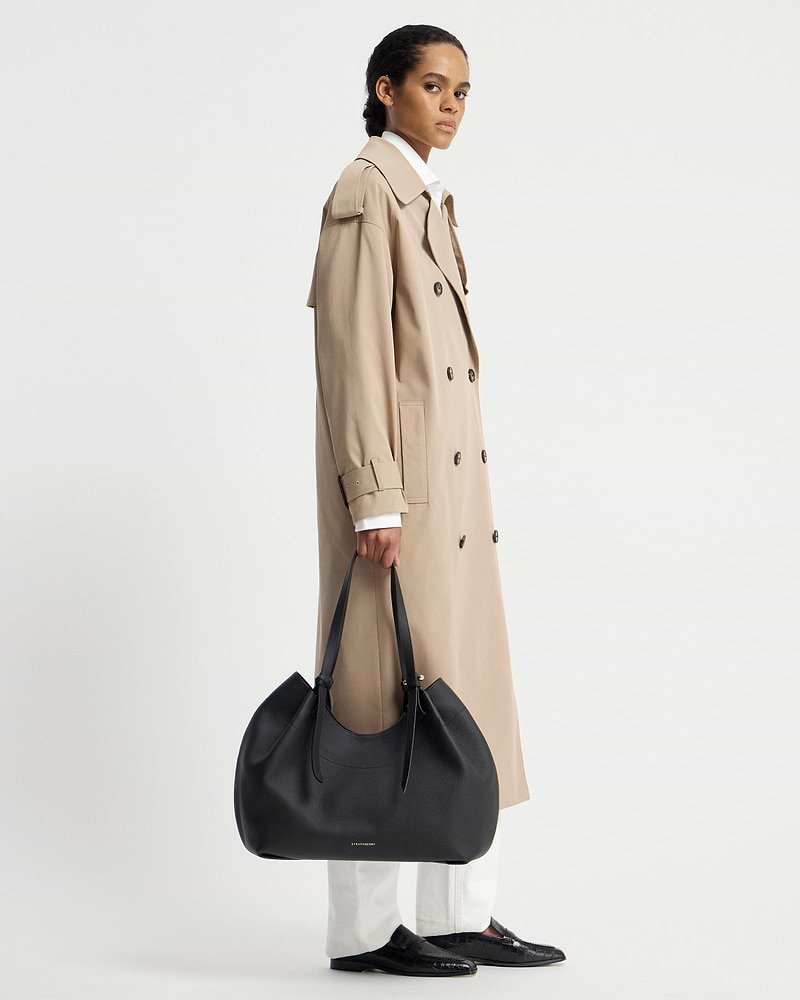 Model in a beige trench coat holding a black Strathberry Barra Tote at her side.
