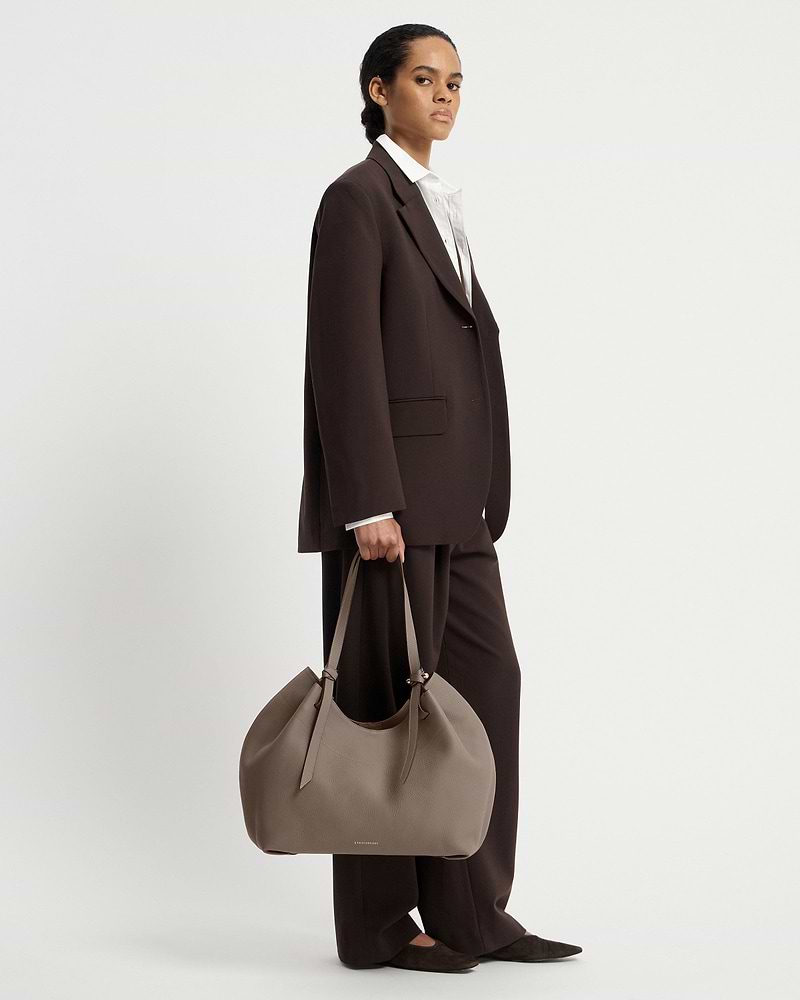 Model in dark suit holding taupe Barra Tote by Strathberry at her side.