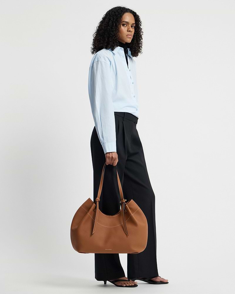 Model wearing light blue shirt holding tan Strathberry Barra tote by her side
