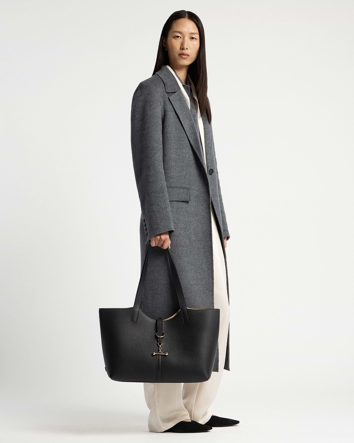 Model in grey coat holding black tote bag with gold hardware by her side.