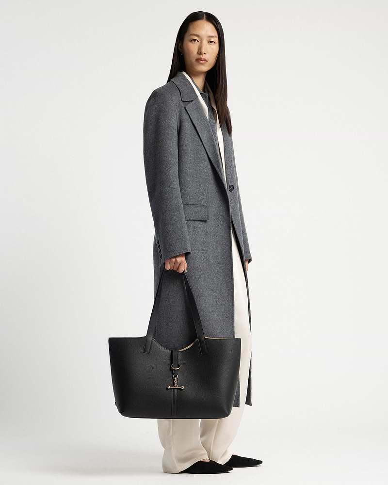 Model in grey coat holding black tote bag with gold hardware by her side.