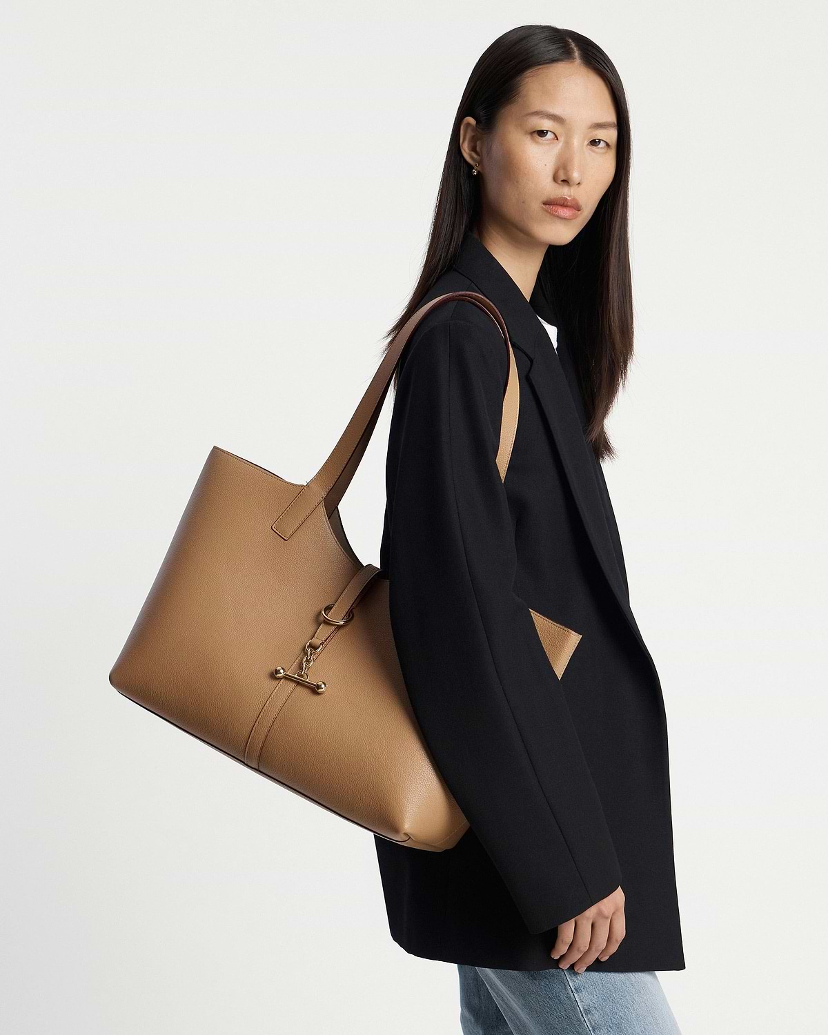 Model wearing black blazer and jeans carrying a caramel leather kite tote on her shoulder.