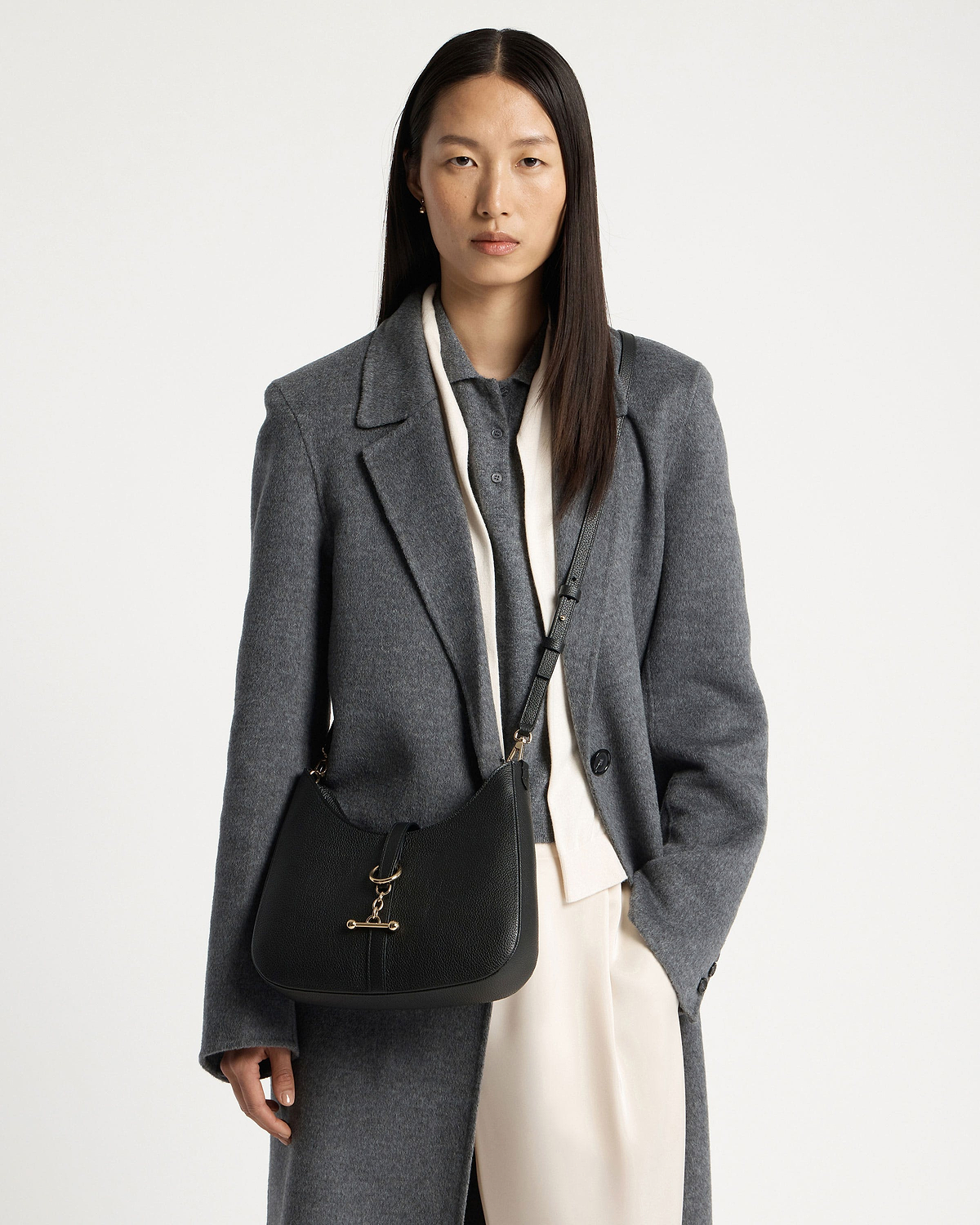 Woman in grey coat wearing black kite sling bag with gold bar fastening.