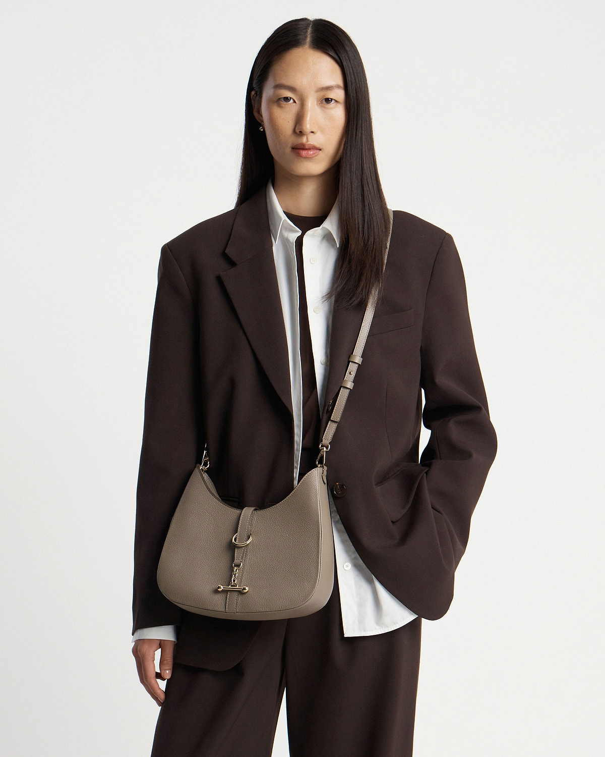 Model wearing taupe kite sling bag cross-body with gold hardware and dark blazer