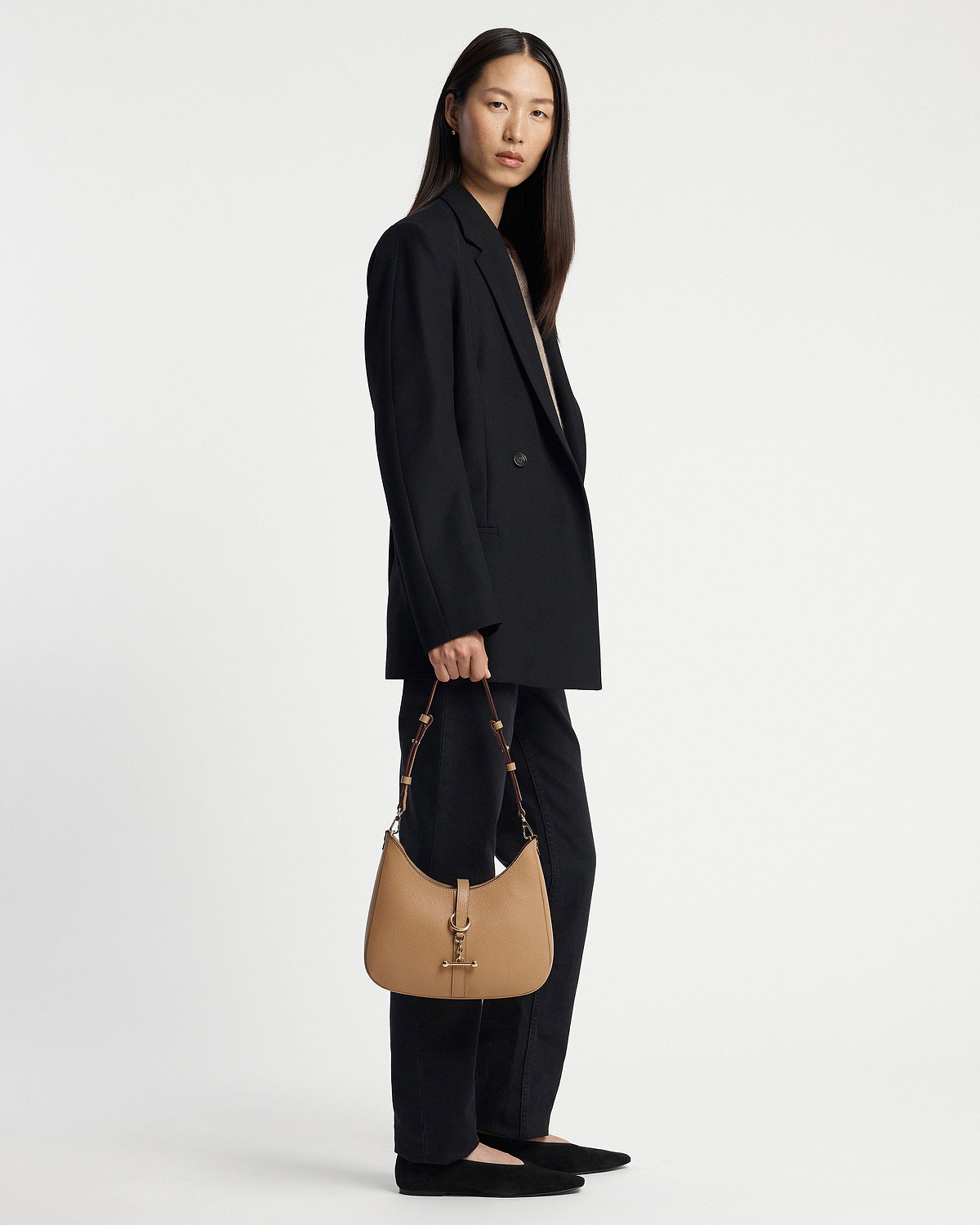Model standing full-length holding a caramel Kite Sling bag by hand.