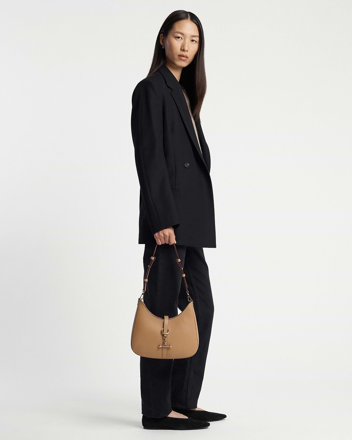 Model standing full-length holding a caramel Kite Sling bag by hand.