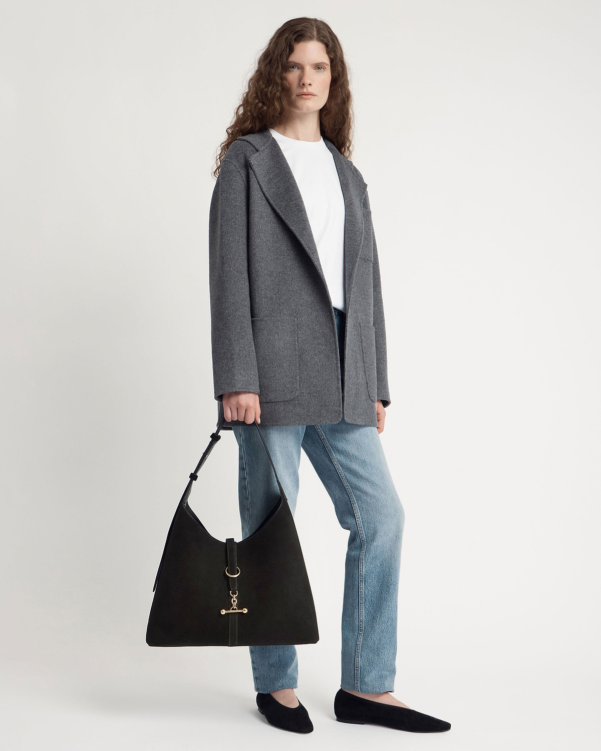 Model wearing grey coat and jeans holding black suede hobo bag.