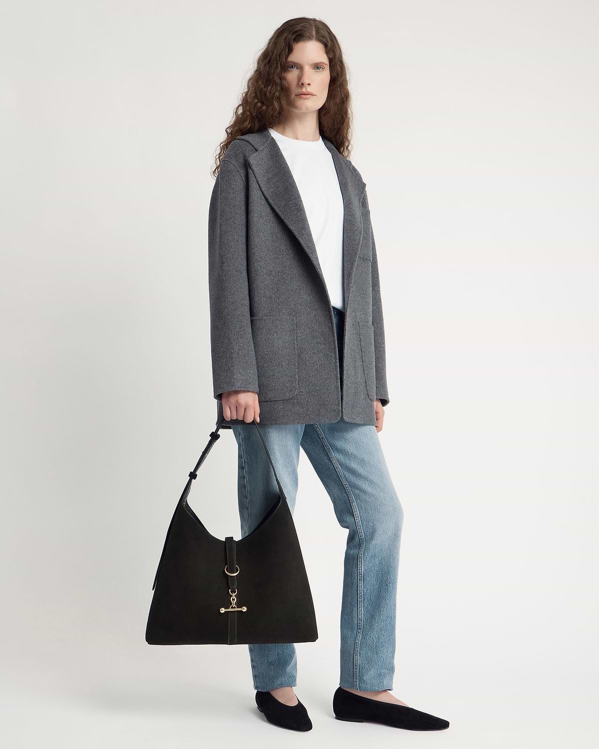 Model wearing grey coat and jeans holding black suede hobo bag.