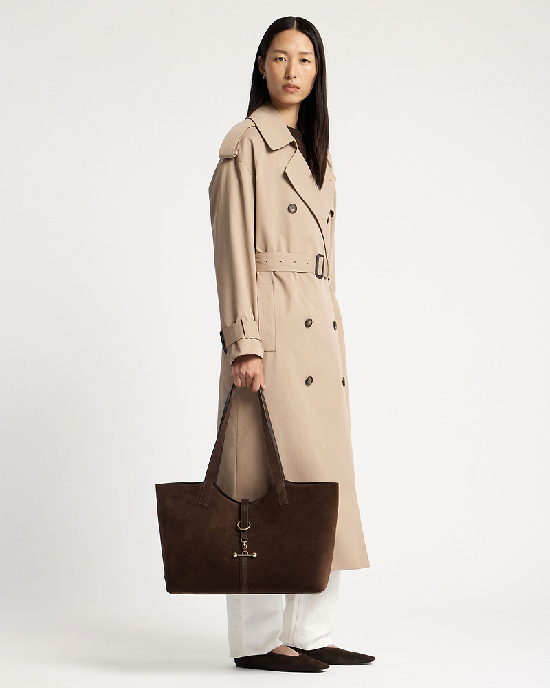Woman in beige trench holding chocolate suede Kite tote with gold hardware.