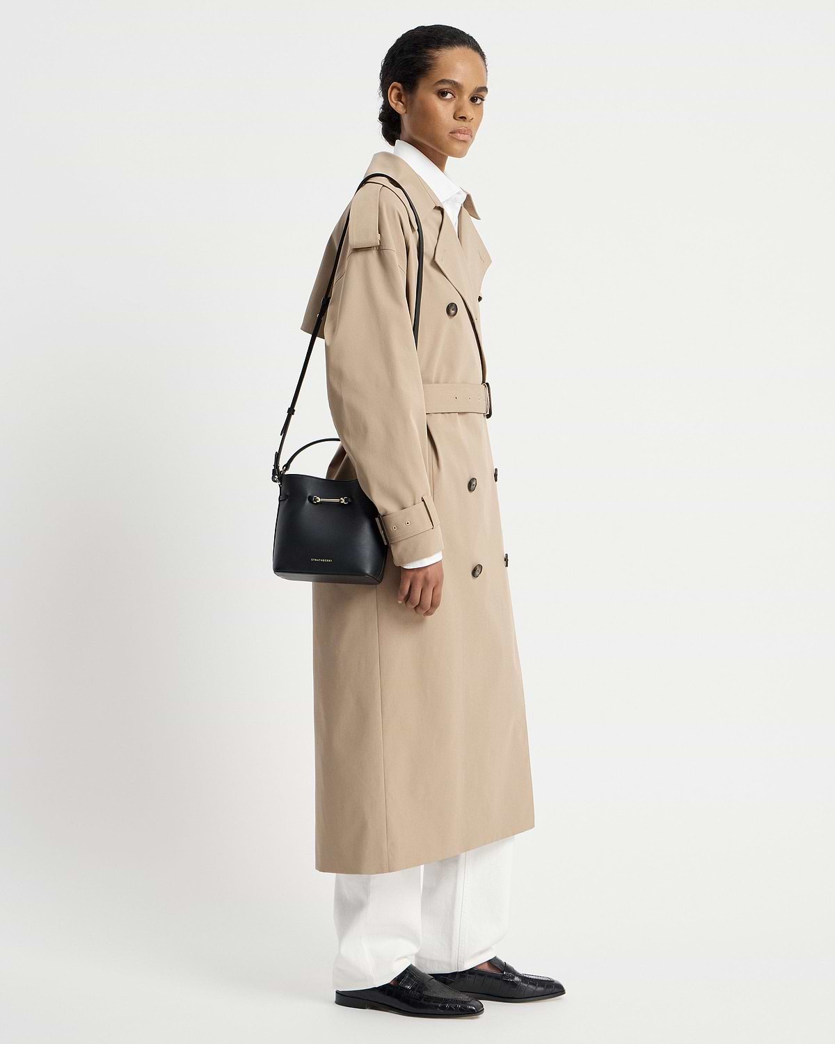 Model in beige trench coat carrying black Strathberry Corda Bucket Mini shoulder bag.
