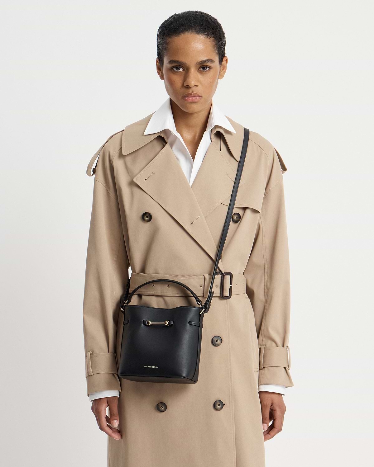 Model wearing a beige trench and black Strathberry Corda Bucket Mini cross-body bag.