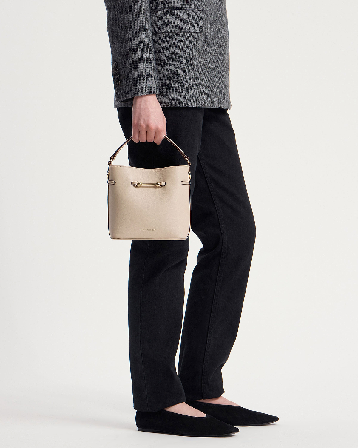 Hand holding an oat‑coloured leather mini bucket bag with top handle