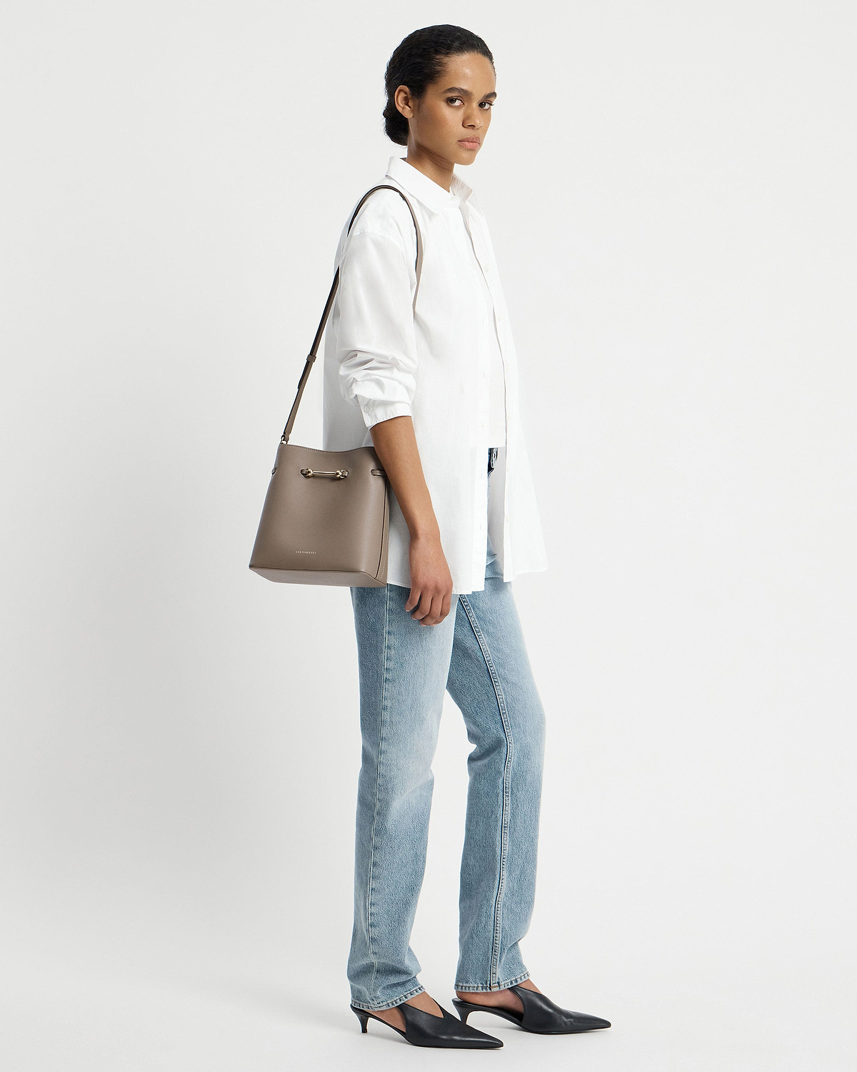 Model wearing a white shirt and jeans, carrying a taupe Strathberry Corda bucket bag.