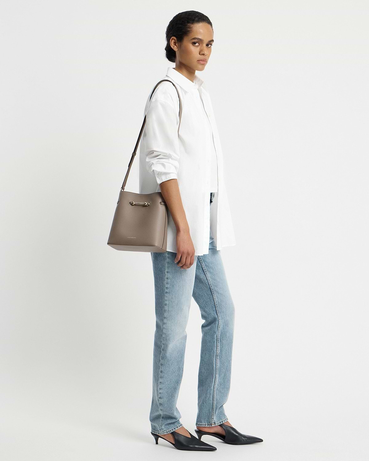 Model wearing a white shirt and jeans, carrying a taupe Strathberry Corda bucket bag.