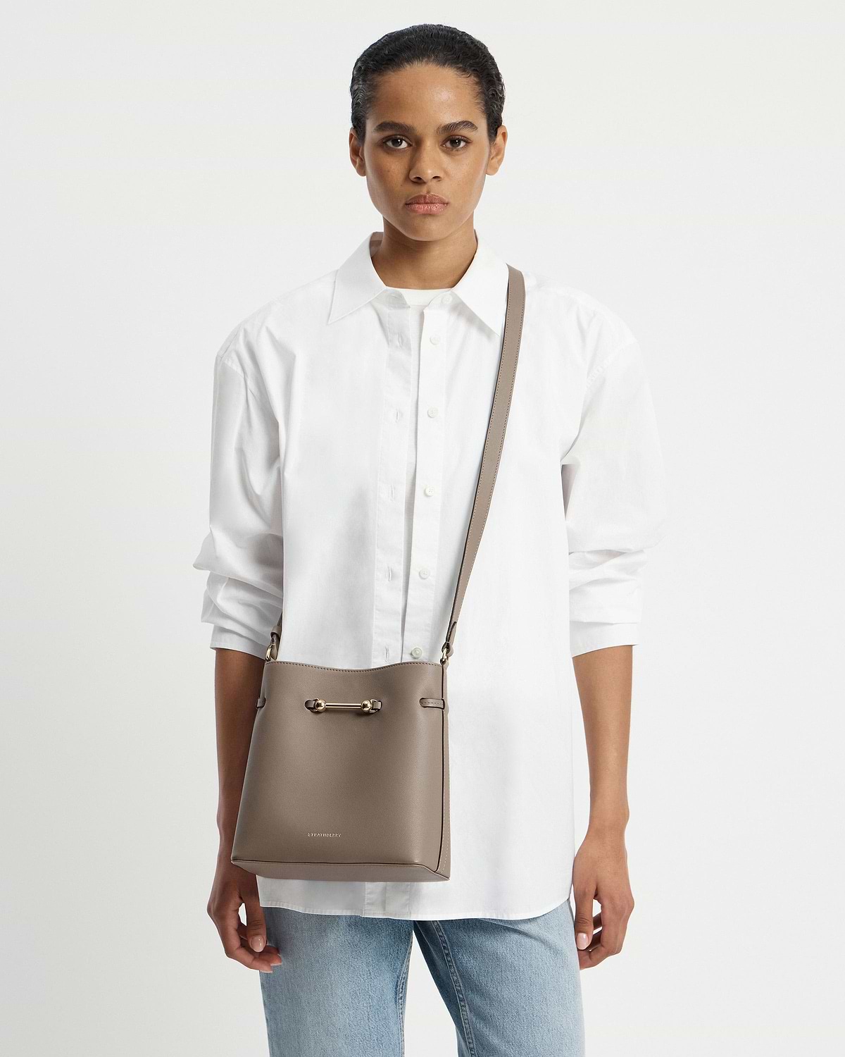 Model wearing white shirt with taupe Corda bucket bag worn crossbody, Strathberry logo visible