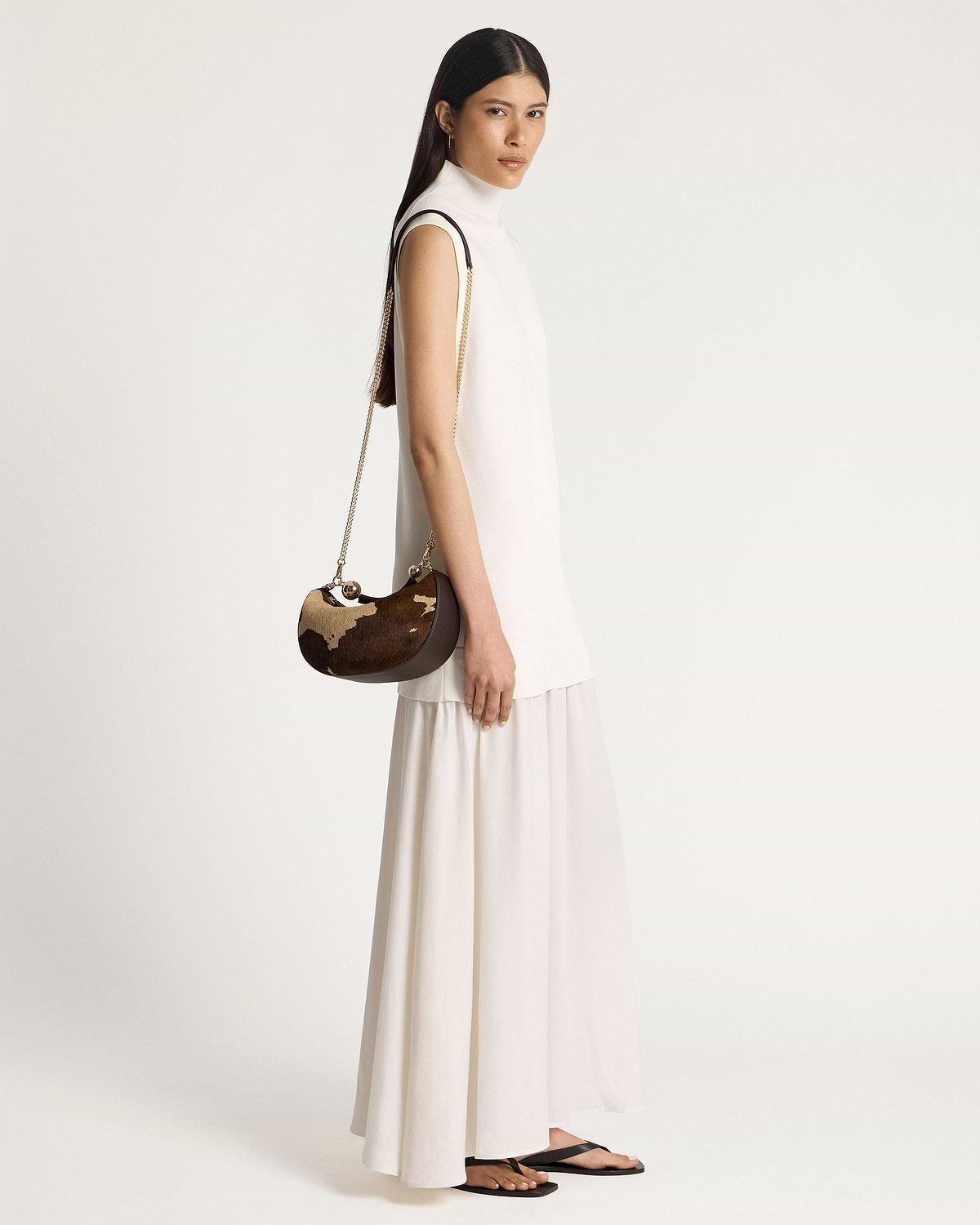 Model in sleeveless white dress carrying a Crescent Moon Mini bag, sand espresso spot print.