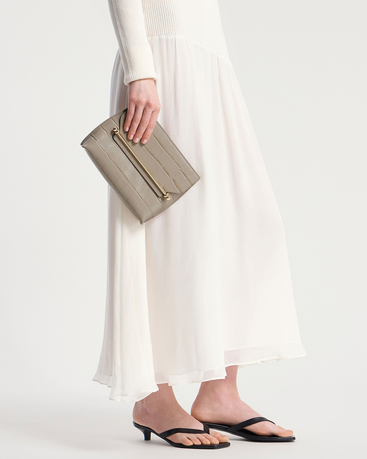 Hand holding croc-embossed clutch with gold bar, model in white dress.