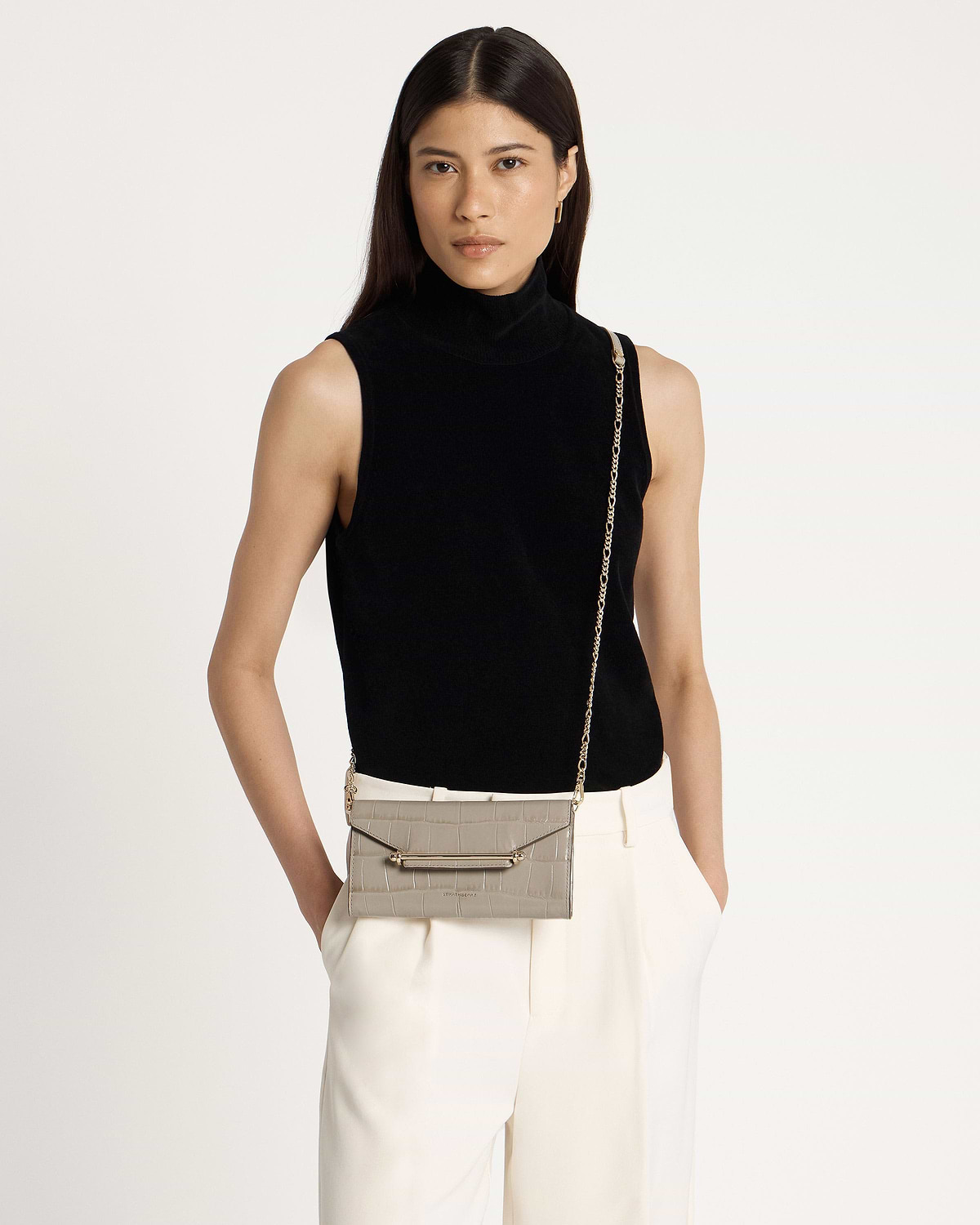 Model in a black sleeveless top and cream trousers with light grey croc-effect chain wallet.