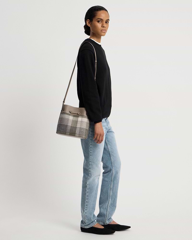 Woman in black jumper and jeans carrying a tartan taupe bucket bag over shoulder.