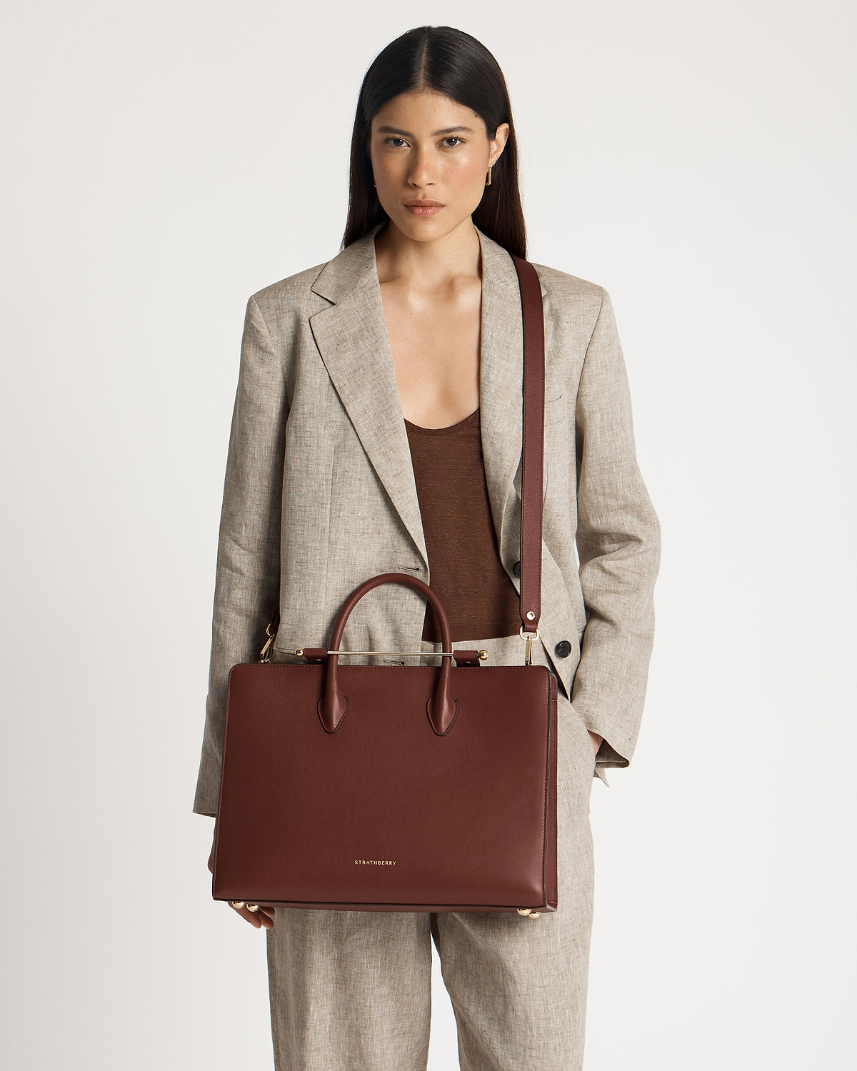 Model in a beige suit holding a walnut Strathberry leather tote bag.