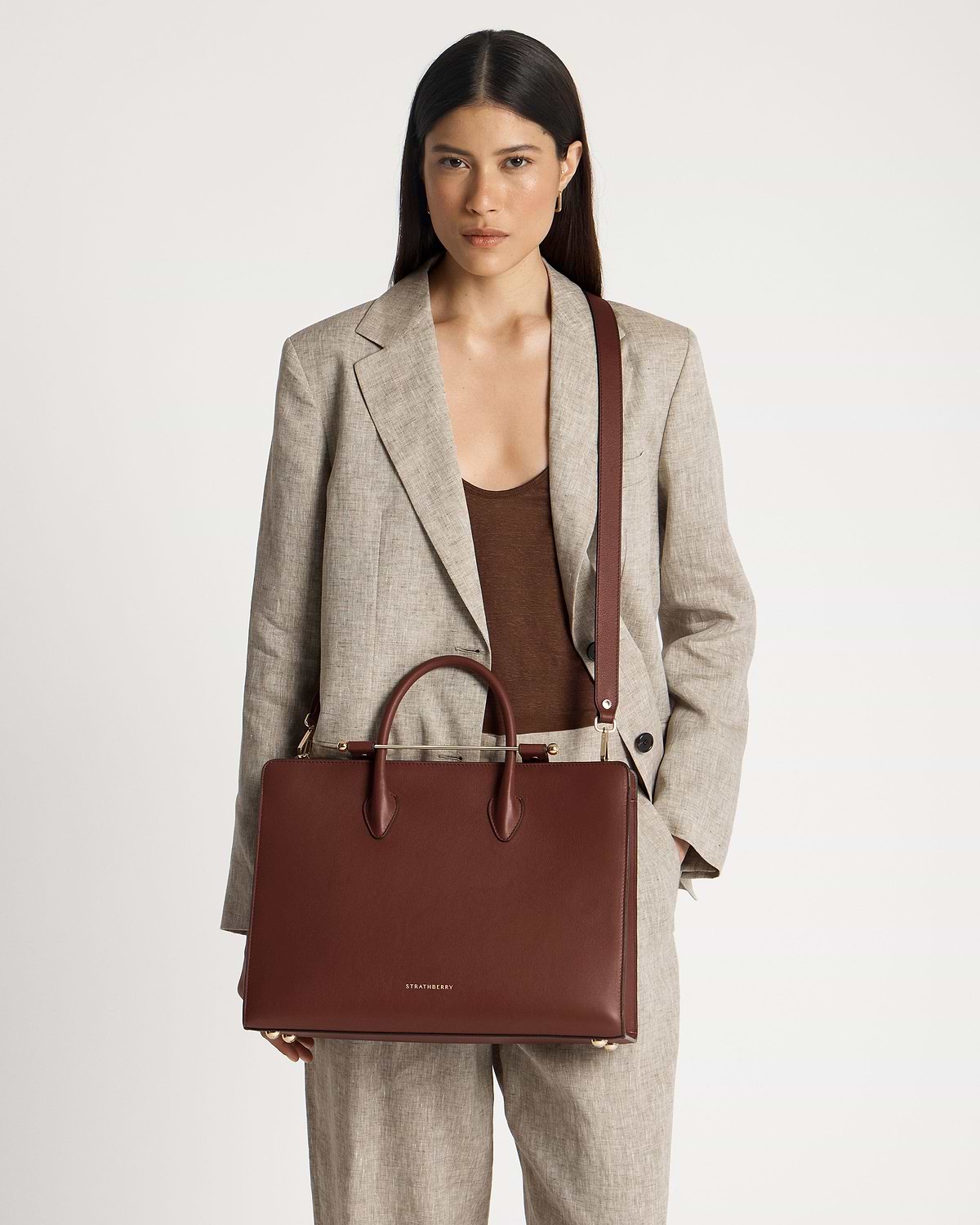 Model in a beige suit holding a walnut Strathberry leather tote bag.