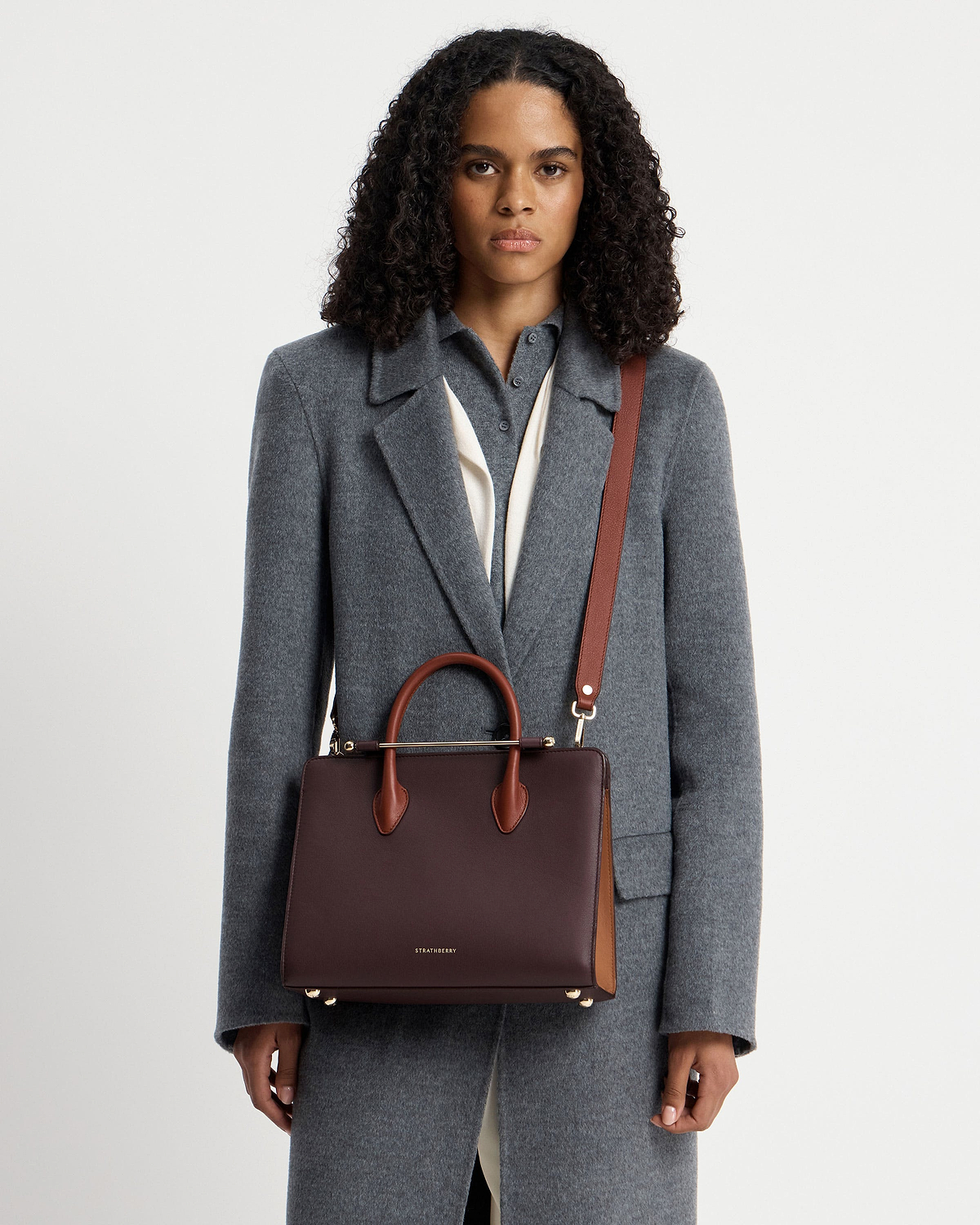 Model wearing grey coat holding Strathberry midi tote in burgundy, walnut and chestnut