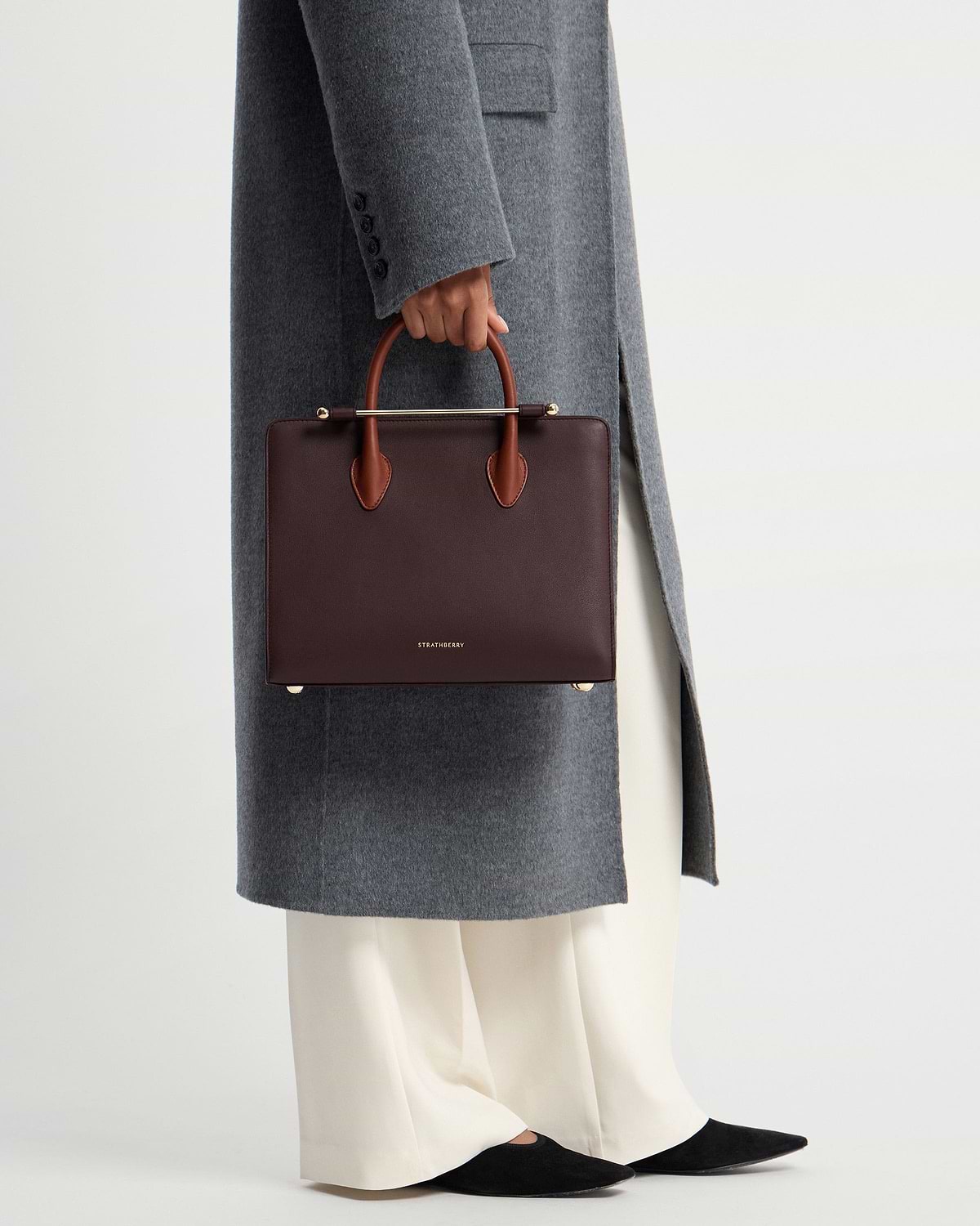 Model waist down holding burgundy Strathberry midi tote with chestnut handles and gold bar.