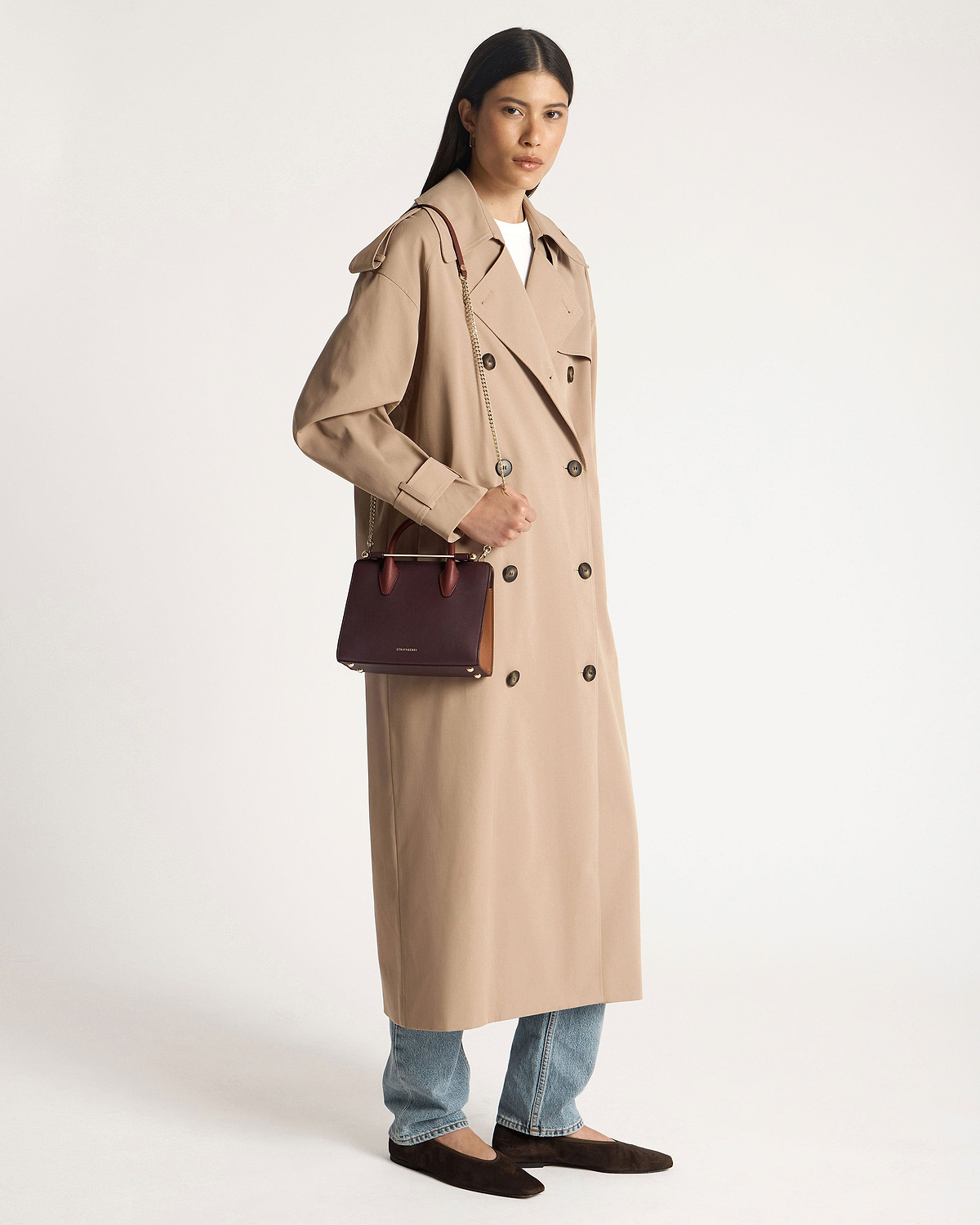 Model wearing beige trench coat holding Strathberry mini tote in burgundy, chestnut, and walnut.