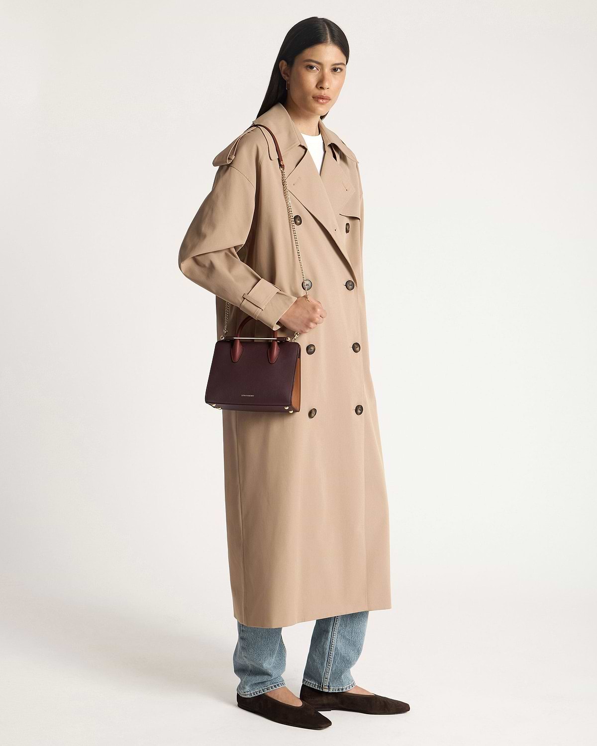 Model wearing beige trench coat holding Strathberry mini tote in burgundy, chestnut, and walnut.