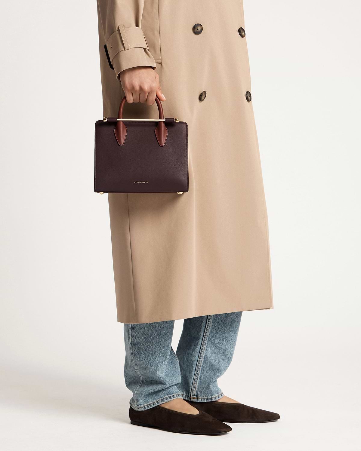 Hand holding Strathberry burgundy mini tote with chestnut handles, model in trench and jeans.