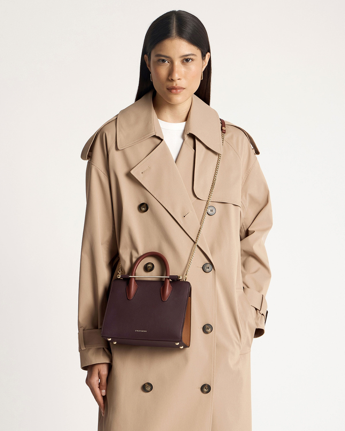 Model wearing beige trench coat with burgundy Strathberry mini tote, chestnut handles, walnut side panel.