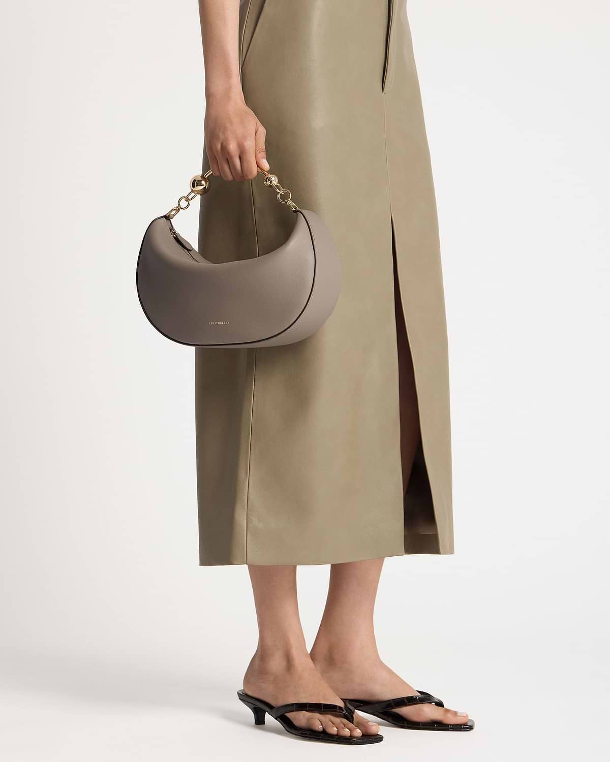 Model holding taupe crescent-shaped handbag with gold chain handle and olive midi skirt.