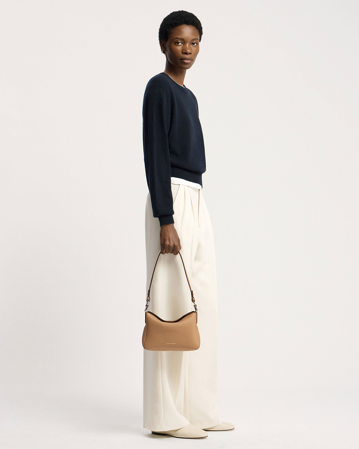 Model holding a caramel Strathberry Sway shoulder bag with top handle and visible logo.