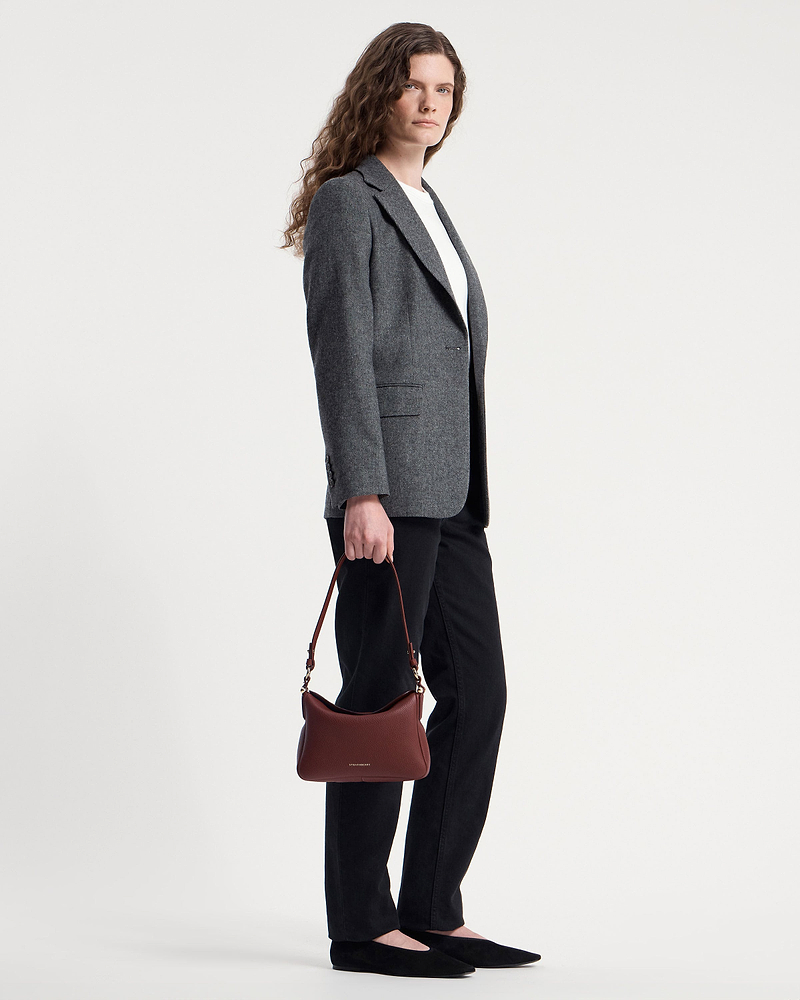 Model holding oxblood Sway shoulder bag by Strathberry while wearing grey blazer.